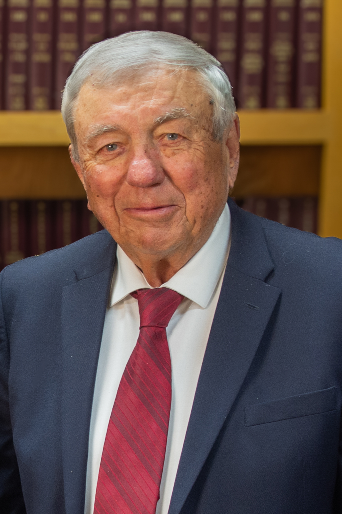 An older man with gray hair, identified as CHARLES P. STEINBAUER, wearing a navy suit, white shirt, and red tie stands in front of shelves filled with books.