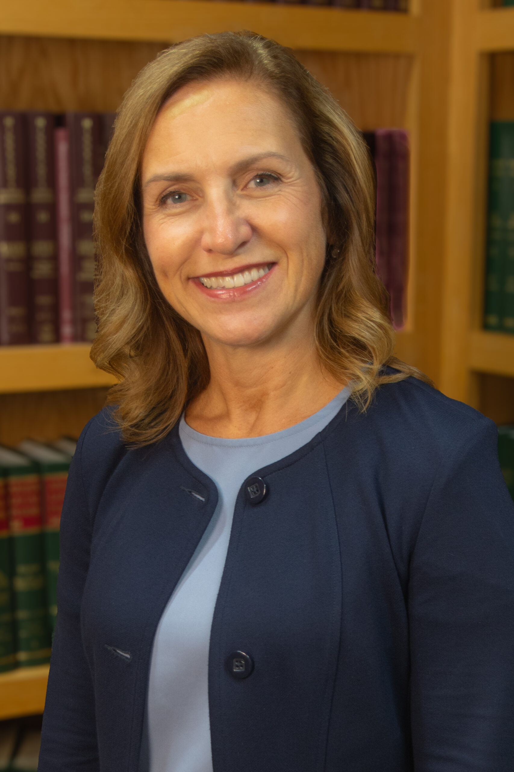 VIRGINIA J. KNUDSON, a woman with shoulder-length blonde hair, wearing a navy blazer over a light blue top, smiles while standing in front of bookshelves filled with books.