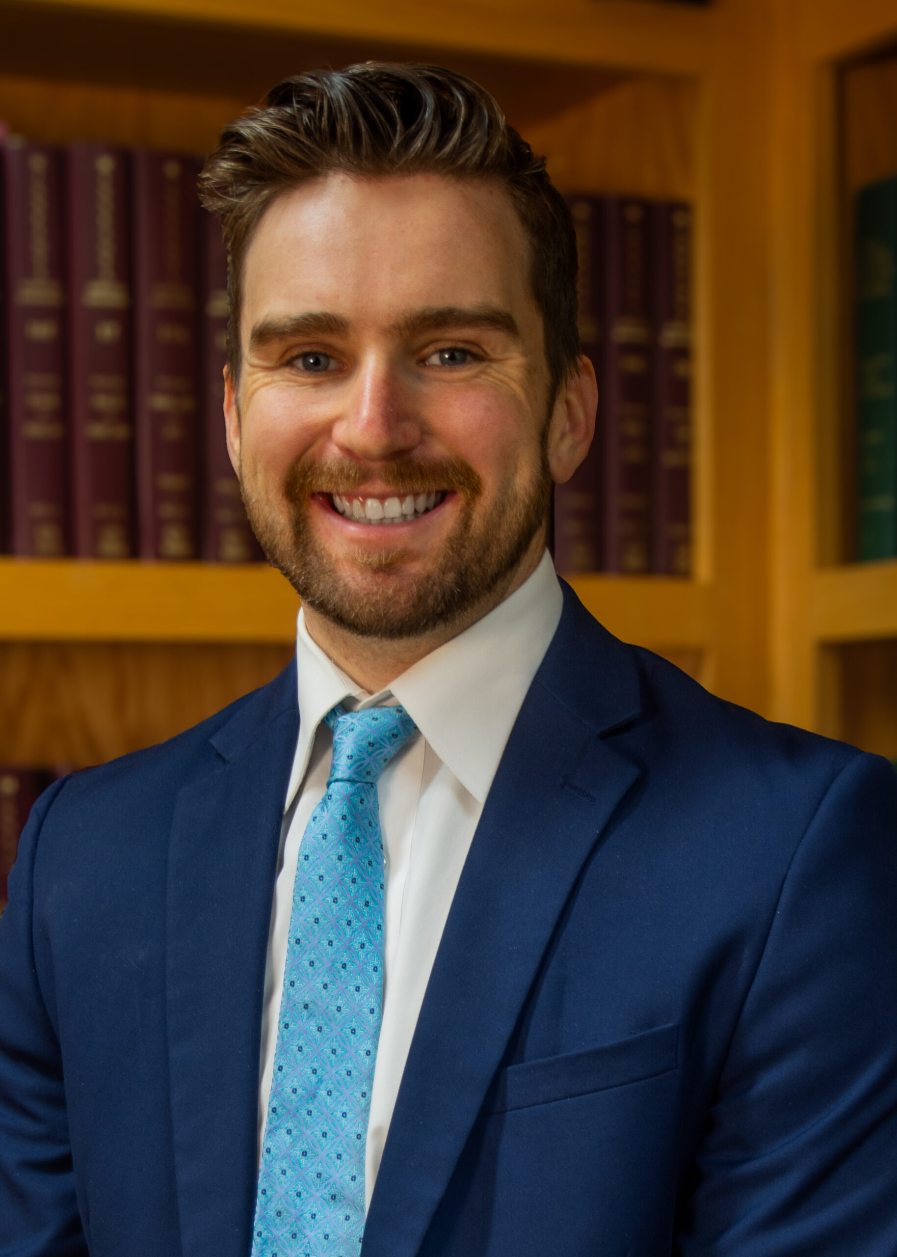 A smiling man, SAMUEL D. CLAUSEN, with short brown hair and a beard, wears a blue suit, white shirt, and light blue patterned tie, standing in front of bookshelves filled with books.