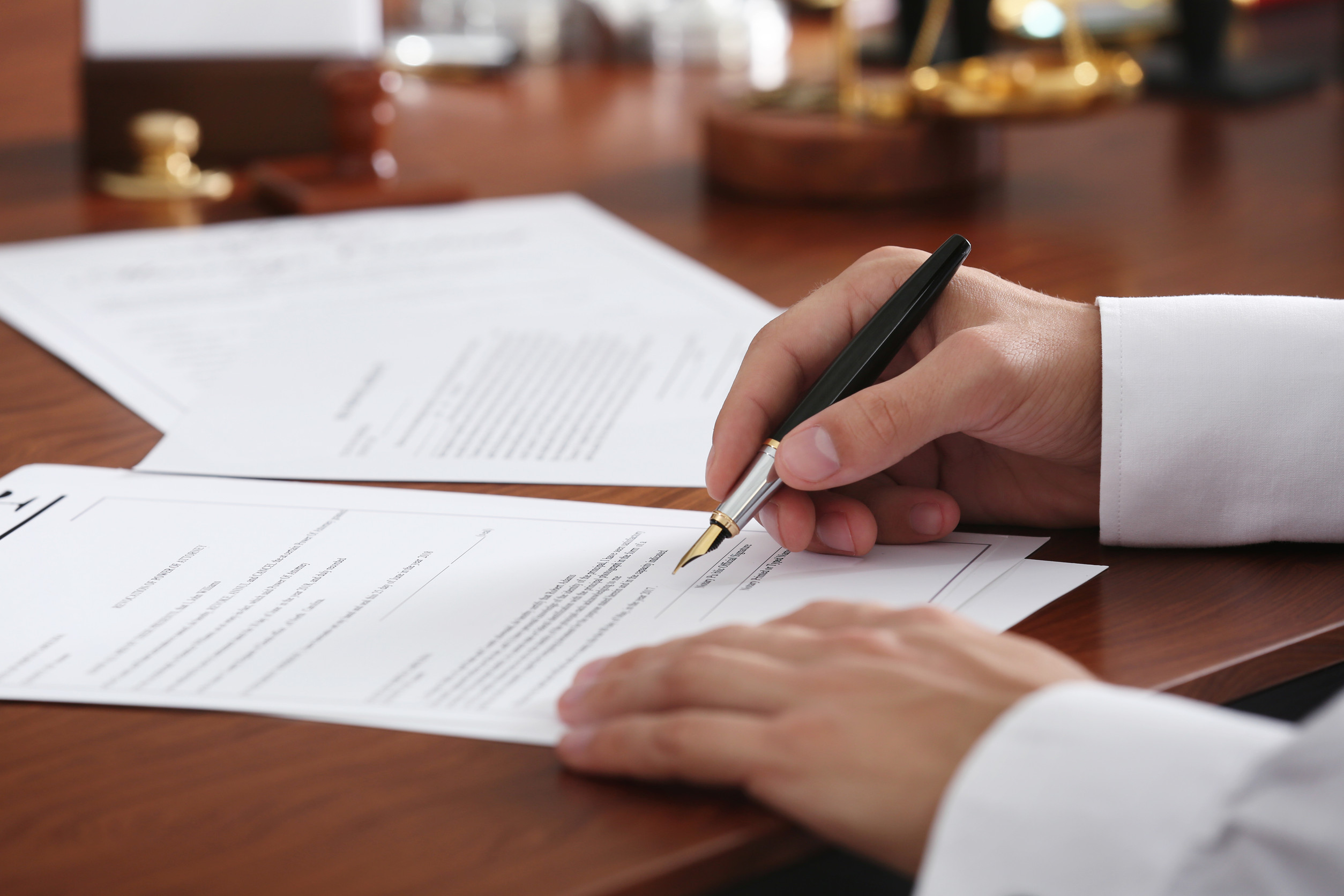 A person in a white shirt is signing an estate planning document with a black and gold pen at a wooden desk, with more papers related to wills and trusts visible among office supplies in the background.