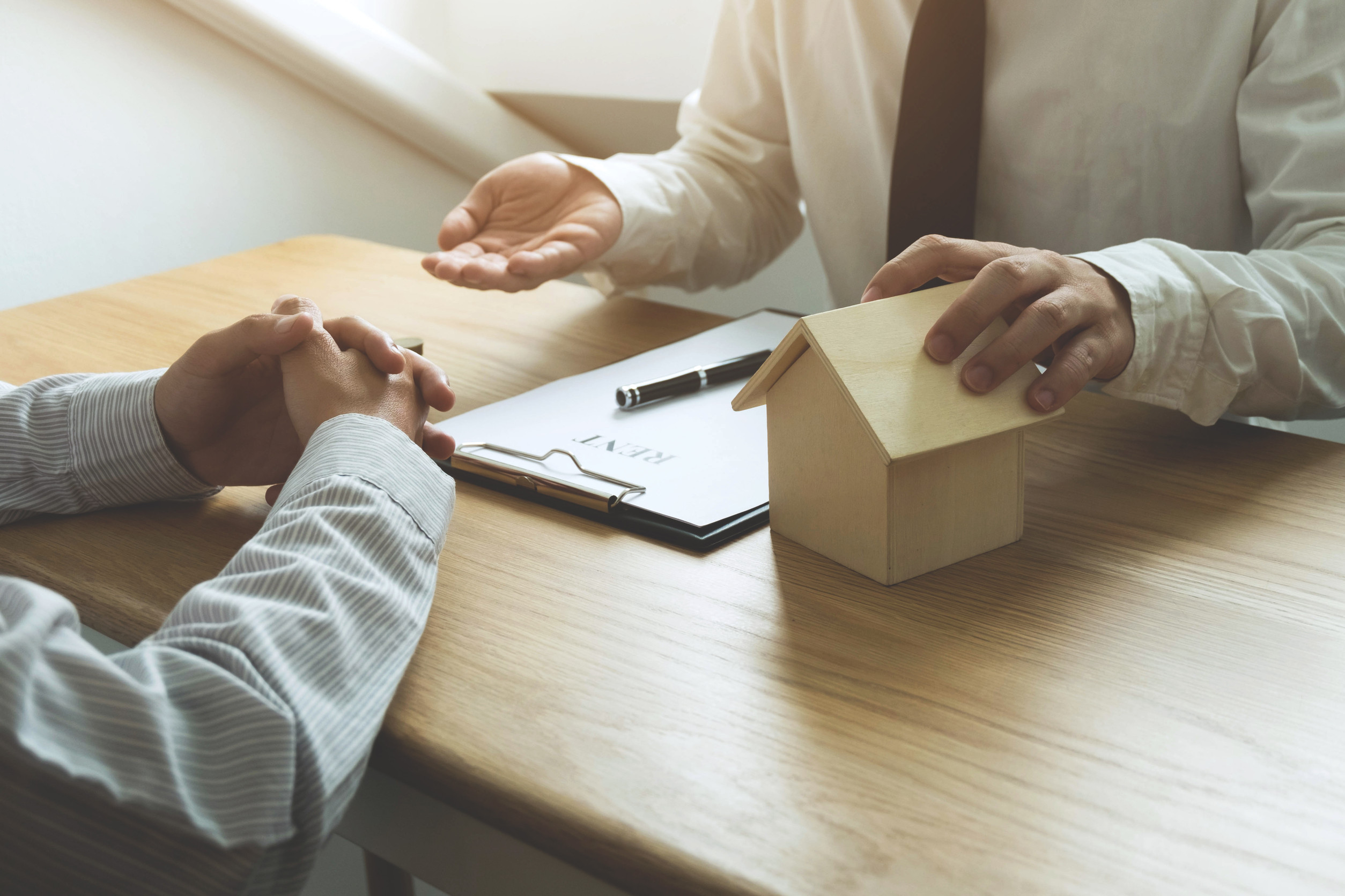 Two people sit at a desk discussing housing or real estate; one holds a small wooden house model while the other has folded hands. A clipboard with a Buy-Sell Agreement labeled "RENT" and a pen are on the table.