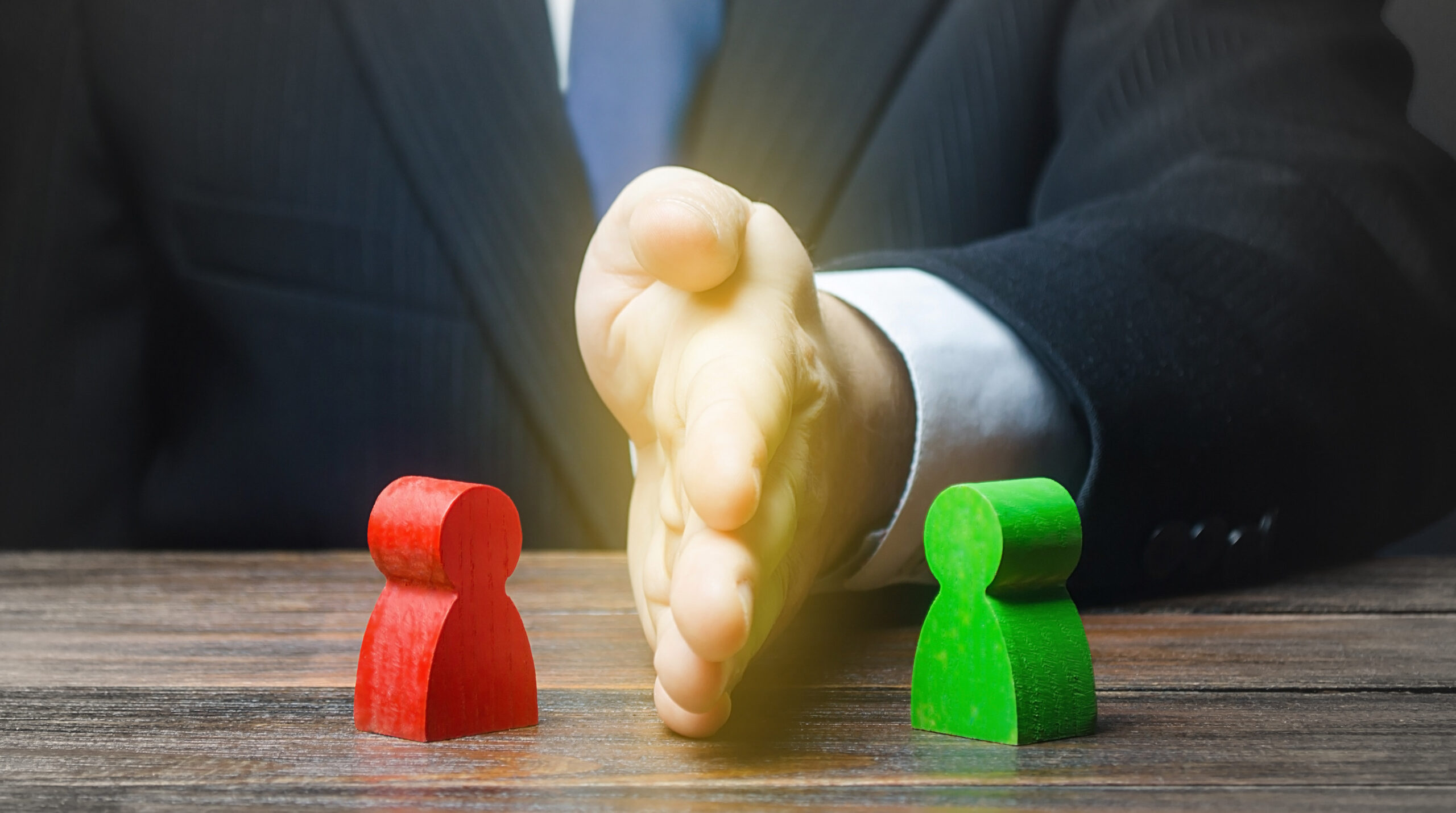 A person in a suit places a hand vertically between two wooden figures, one red and one green, symbolizing mediation or separation of differences on a wooden table.
