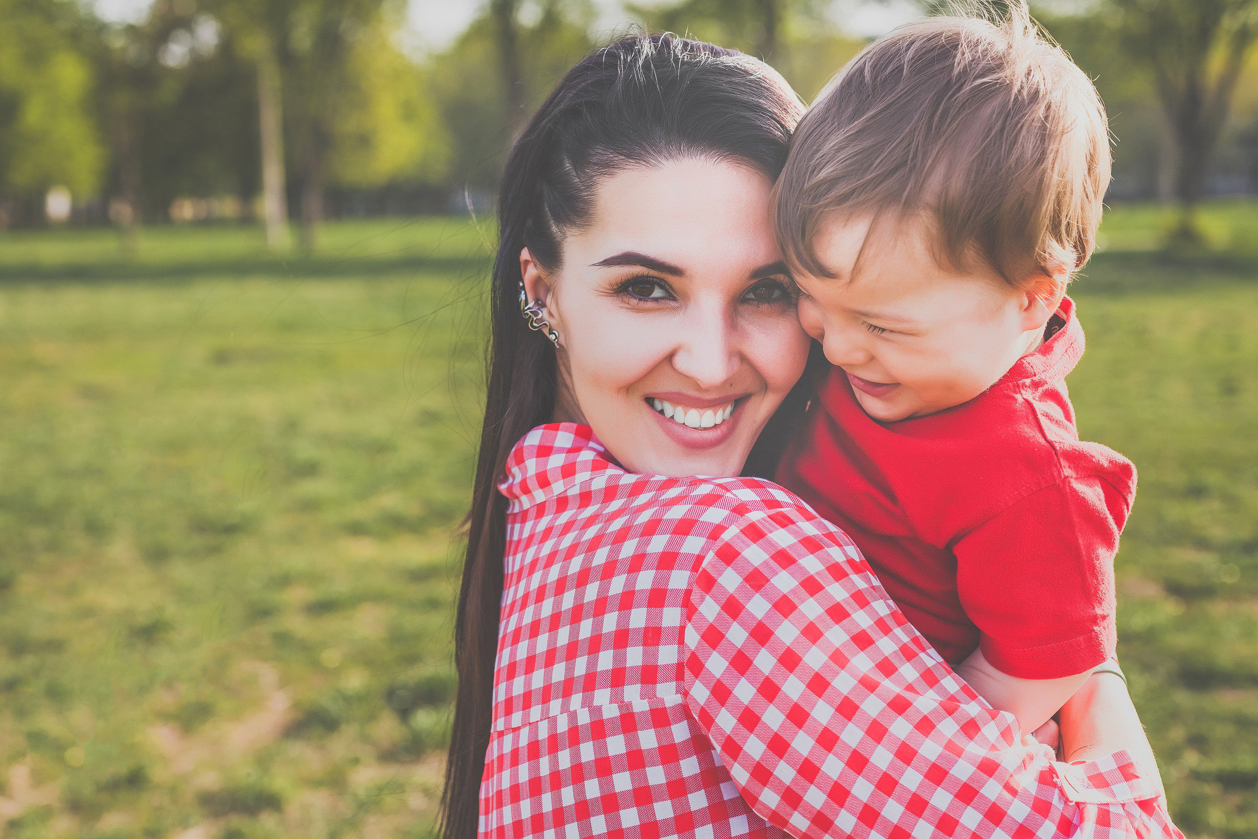 A smiling woman in a red checkered shirt, symbolizing guardianship, holds a laughing young boy dressed in red, outdoors in a grassy park with trees in the background.