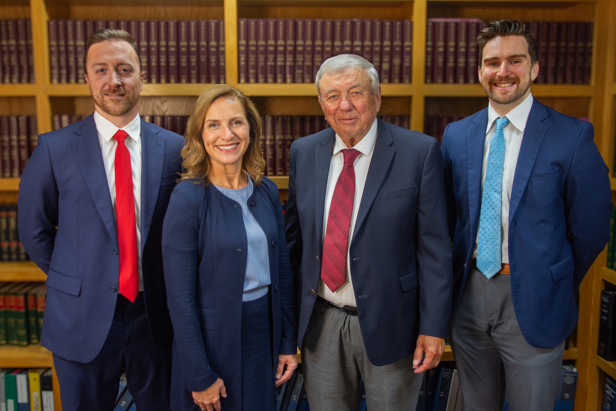 Four people in business attire stand smiling in front of a bookshelf filled with legal books. Three men and one woman are arranged side by side, facing the camera in a professional setting.