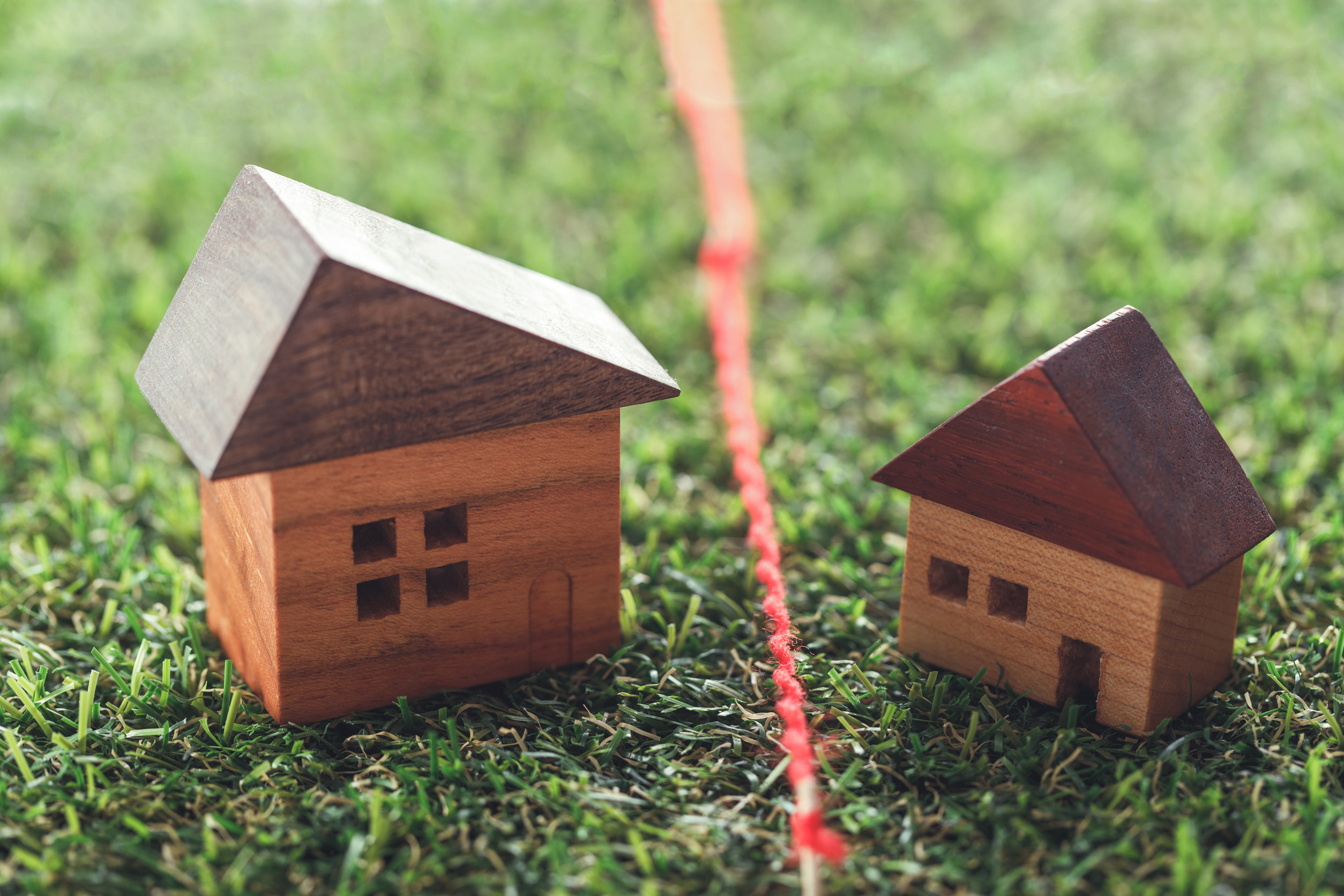 Two small wooden house models sit on grass, separated by a red string line, symbolizing property lines or potential boundary disputes.