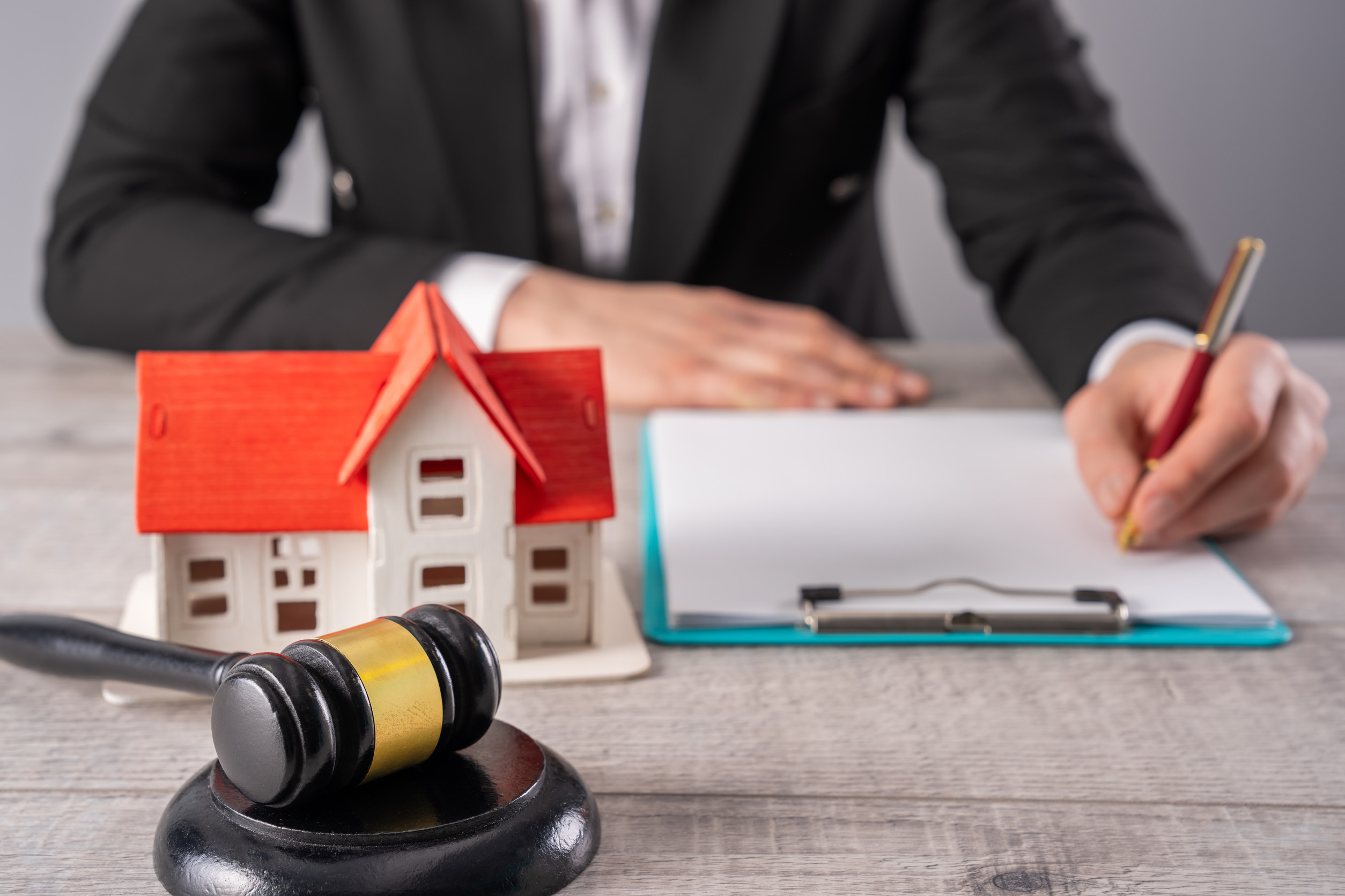 A person in a suit writes on a clipboard at a desk with a small model house and a judge’s gavel, symbolizing real estate proceedings and the use of legal real estate tools such as a 1031 exchange.