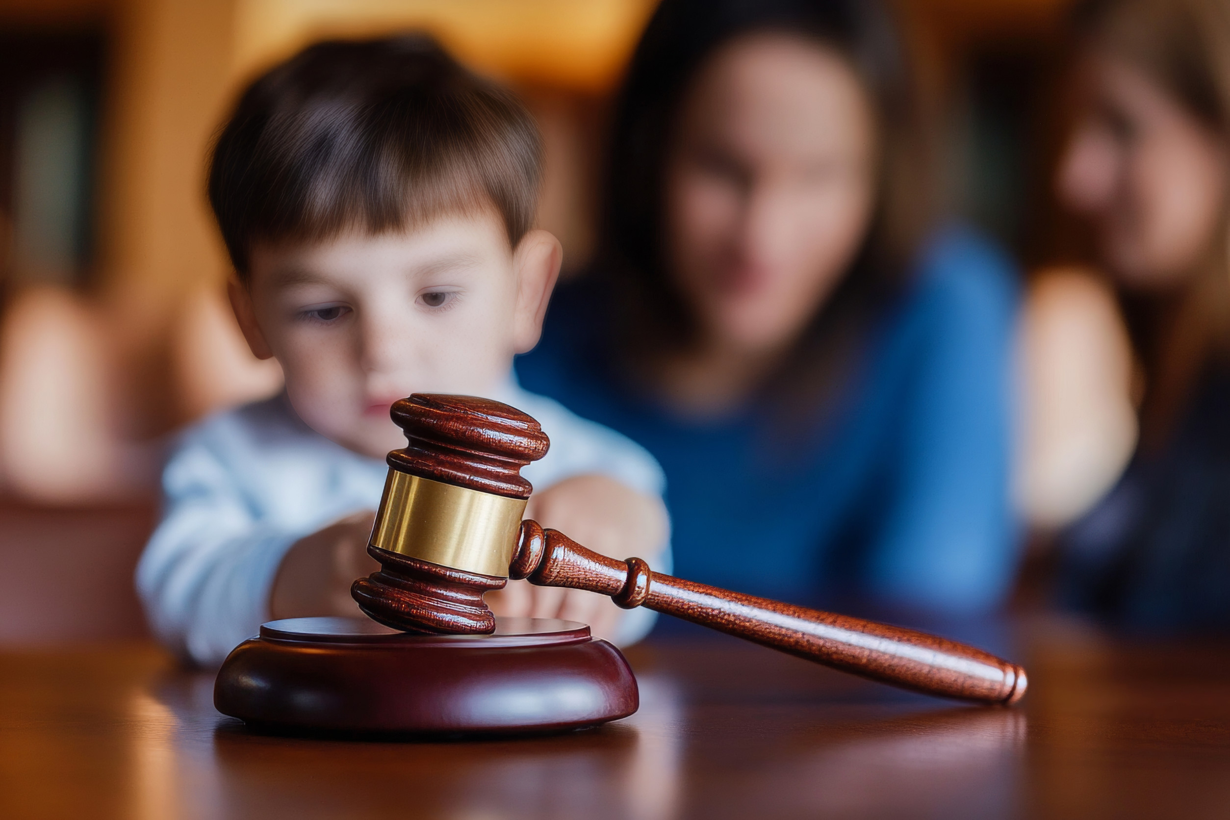 A young child holding a judge’s gavel, with two adults blurred in the background. The child appears curious and focused on the gavel, symbolizing the difference that custody or parenting time decisions can make at a wooden table.