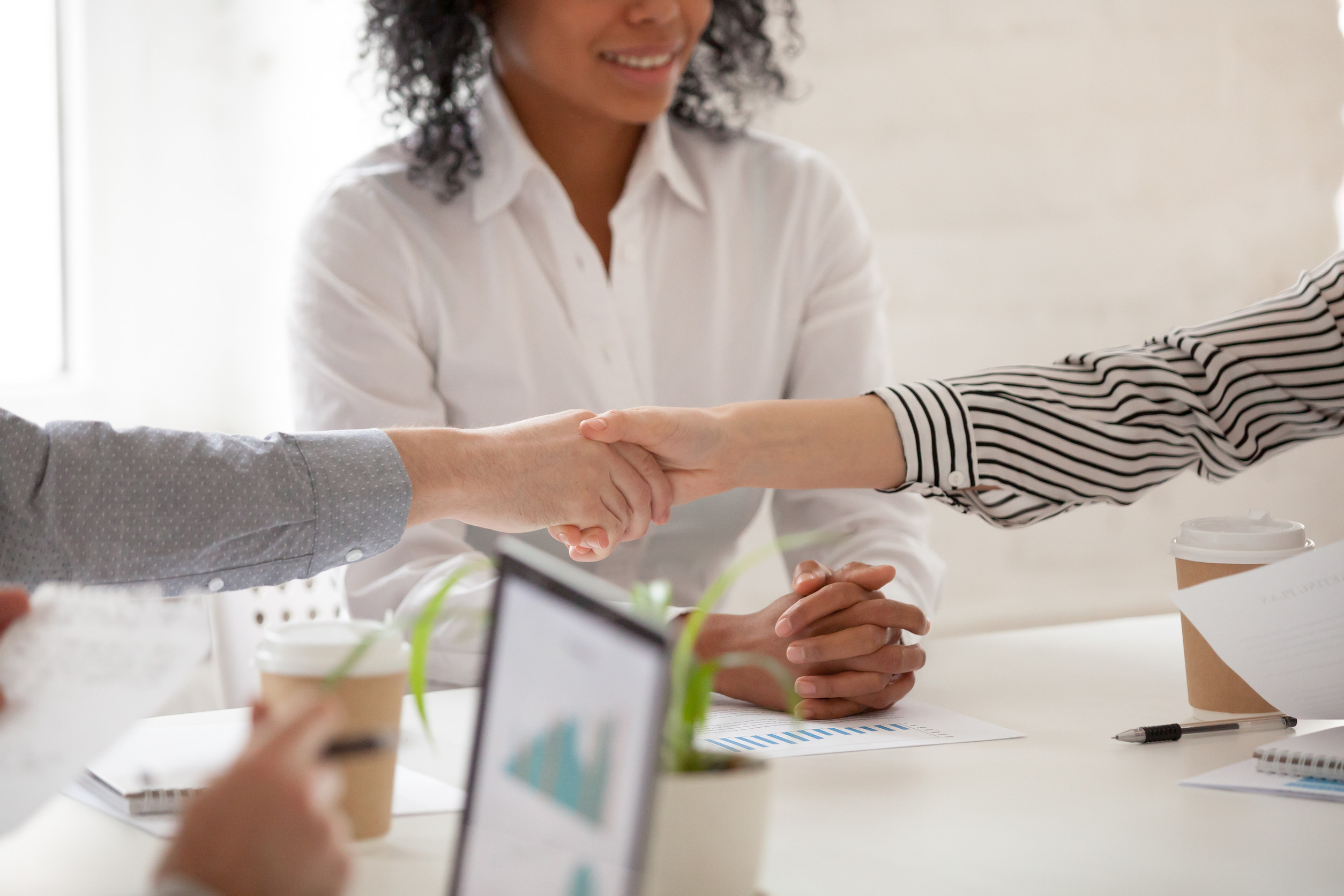 Close-up of two people shaking hands across a table in an office setting, with a woman smiling in the background—highlighting her mediator role amid documents, coffee cups, and a laptop.