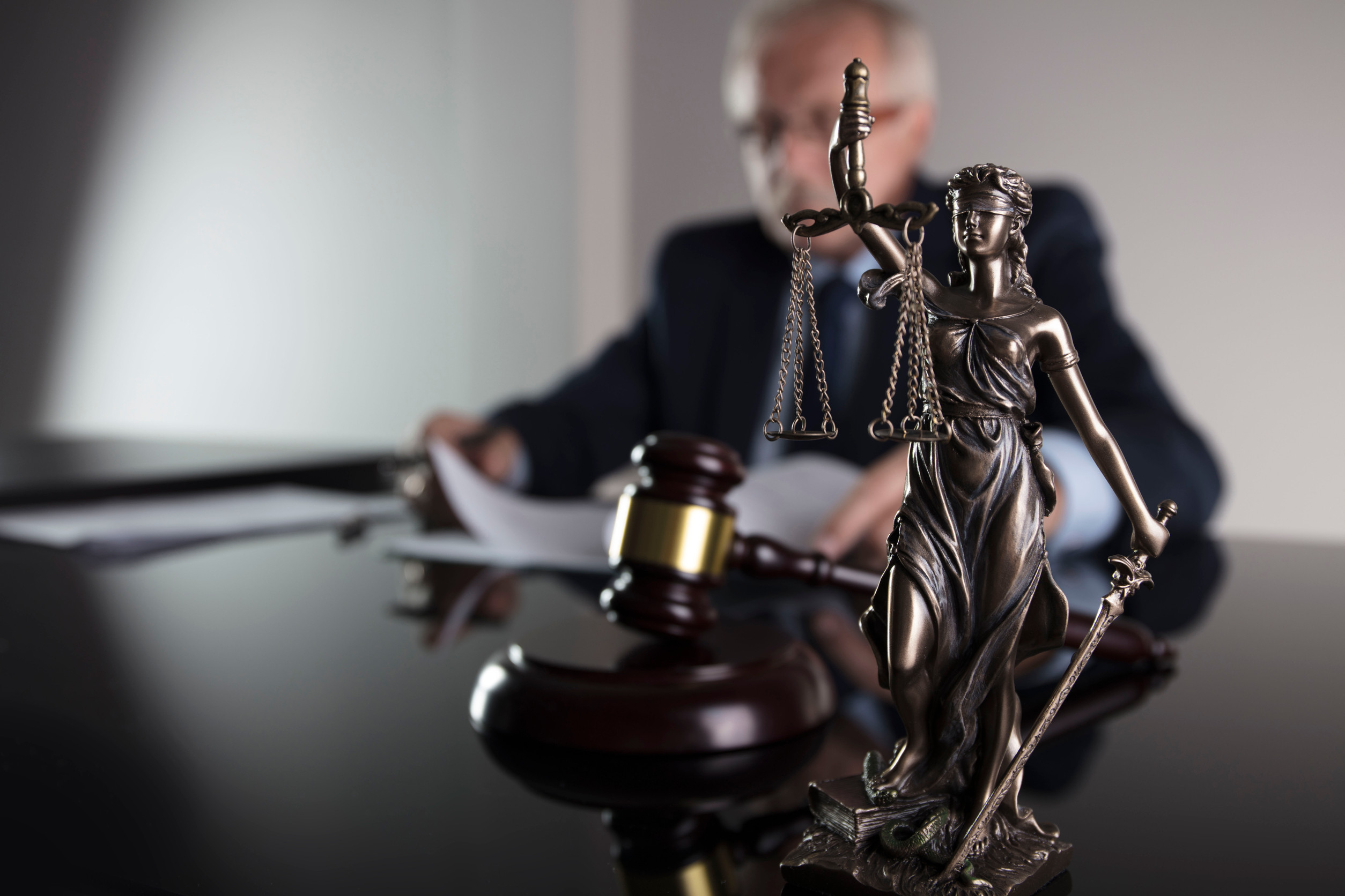 A statue of Lady Justice and a judge’s gavel are on a desk, with a blurred person in a suit reading documents in the background, symbolizing law and justice in everyday legal issues faced by local government.