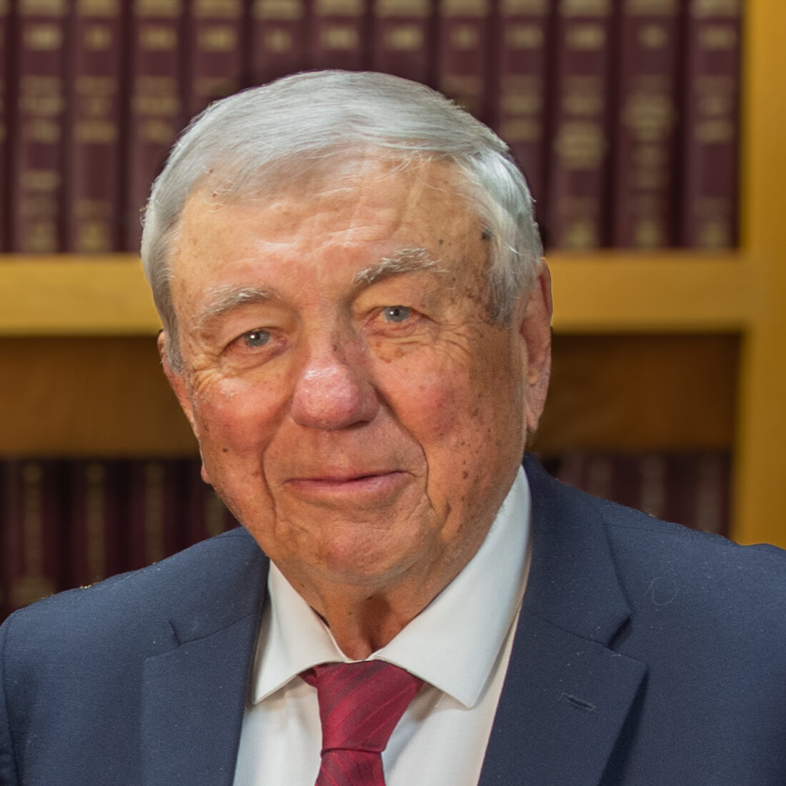 An older man with gray hair in a navy suit, white shirt, and red tie stands in front of bookshelves filled with burgundy- and gold-bound books. He is smiling slightly and looking at the camera.