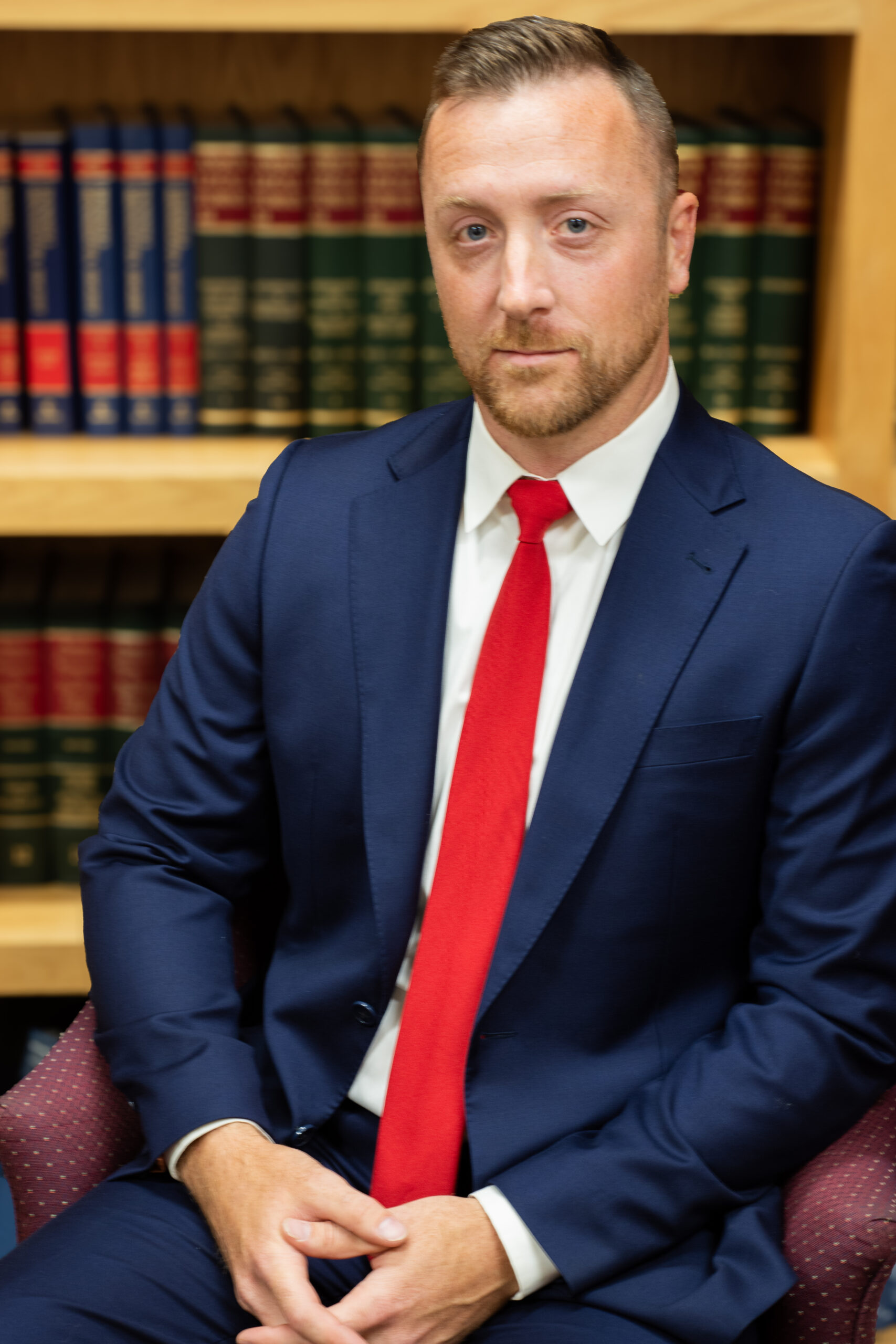 A man in a navy suit and red tie sits in a maroon chair, hands folded, in front of bookshelves filled with legal books.