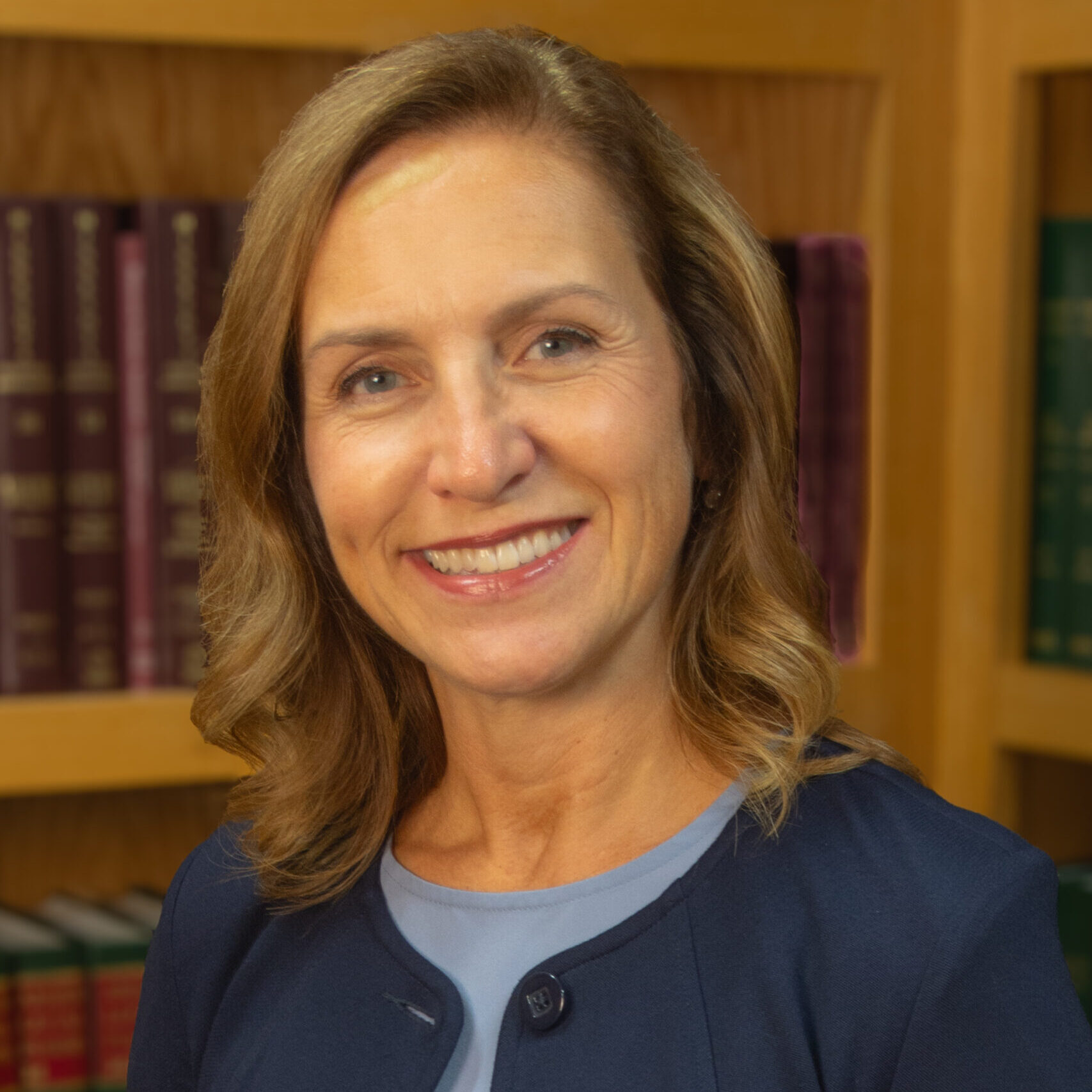 A woman with shoulder-length blonde hair wearing a navy blazer and light blue top smiles in front of bookshelves filled with books.