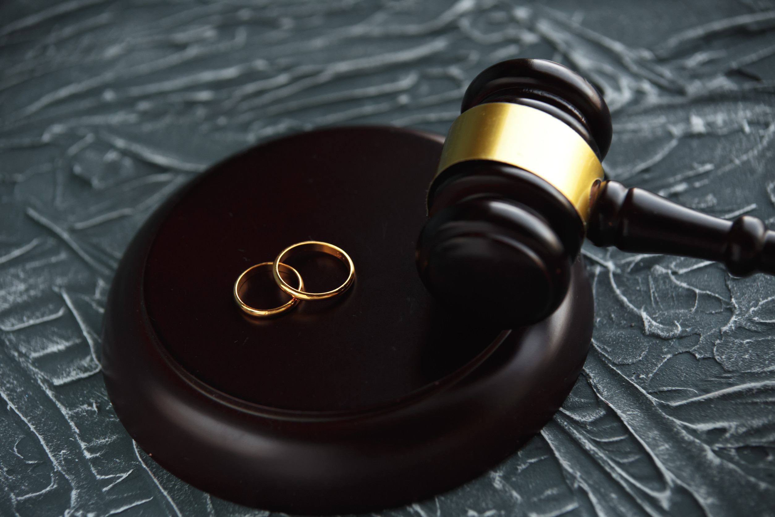 Two gold wedding rings rest on a wooden judge’s gavel and sound block, symbolizing marriage, prenuptial agreements, or divorce on a textured dark background.
