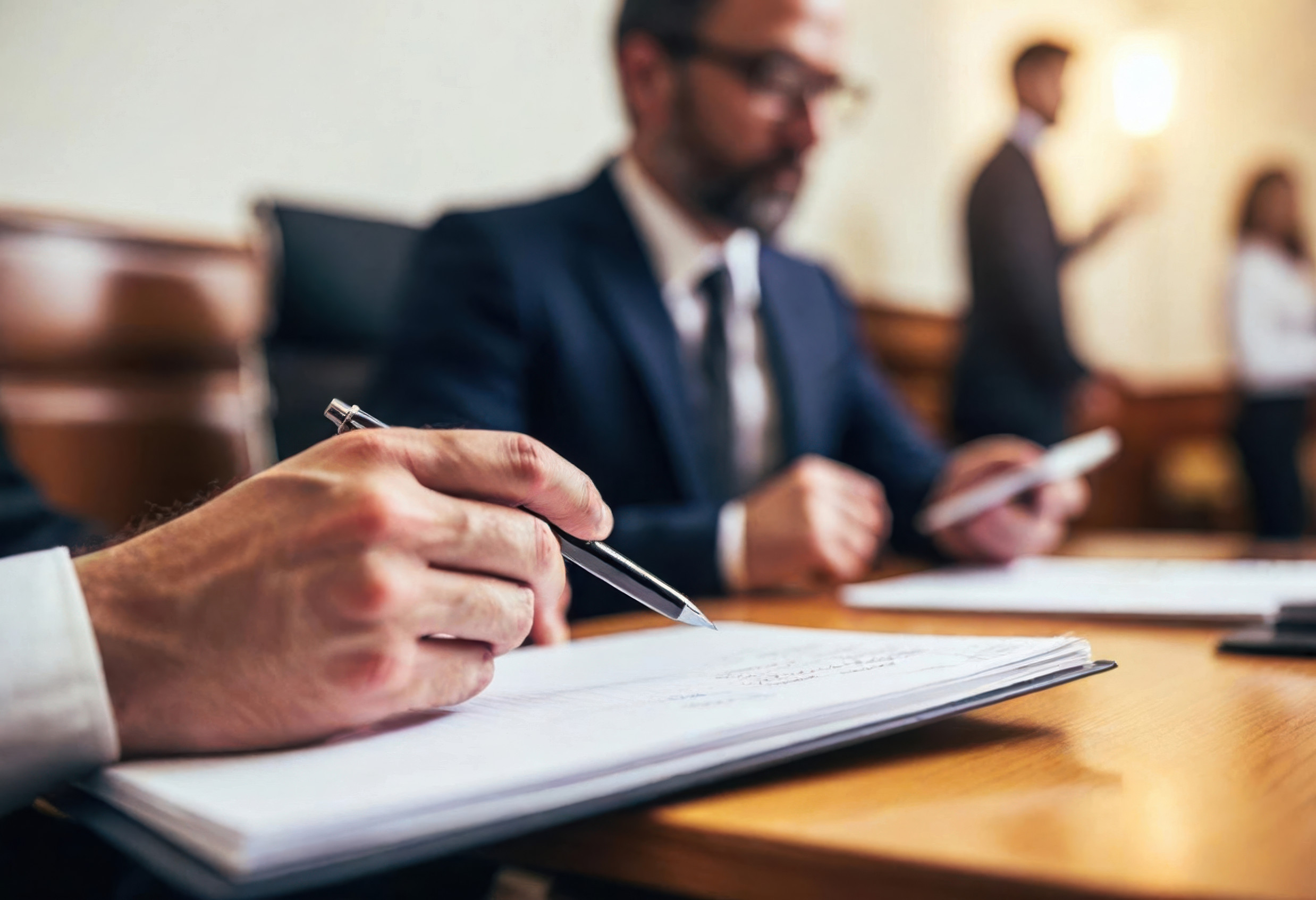 Close-up of a person’s hand holding a pen and writing on documents at a desk in a courtroom, seeking legal counsel, with another person in a suit seated and blurred figures in the background.