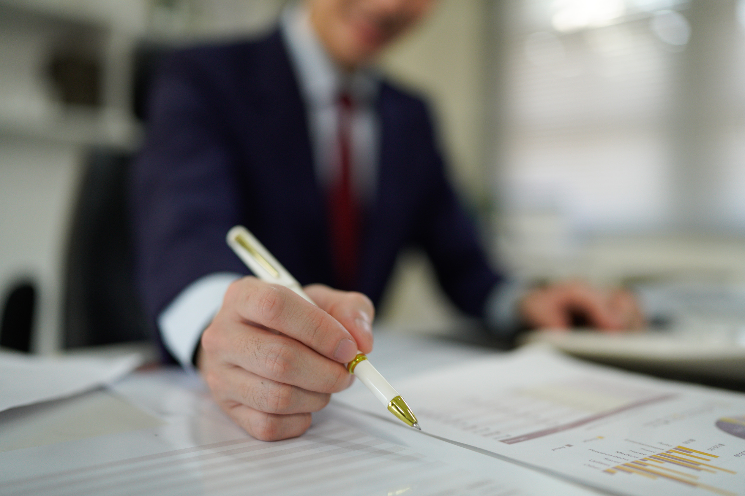 A person in a suit is sitting at a desk, holding a pen and pointing at a document with charts and graphs—perfect for someone researching starting a business in Minnesota. The focus is on the hand and pen, with the face blurred.