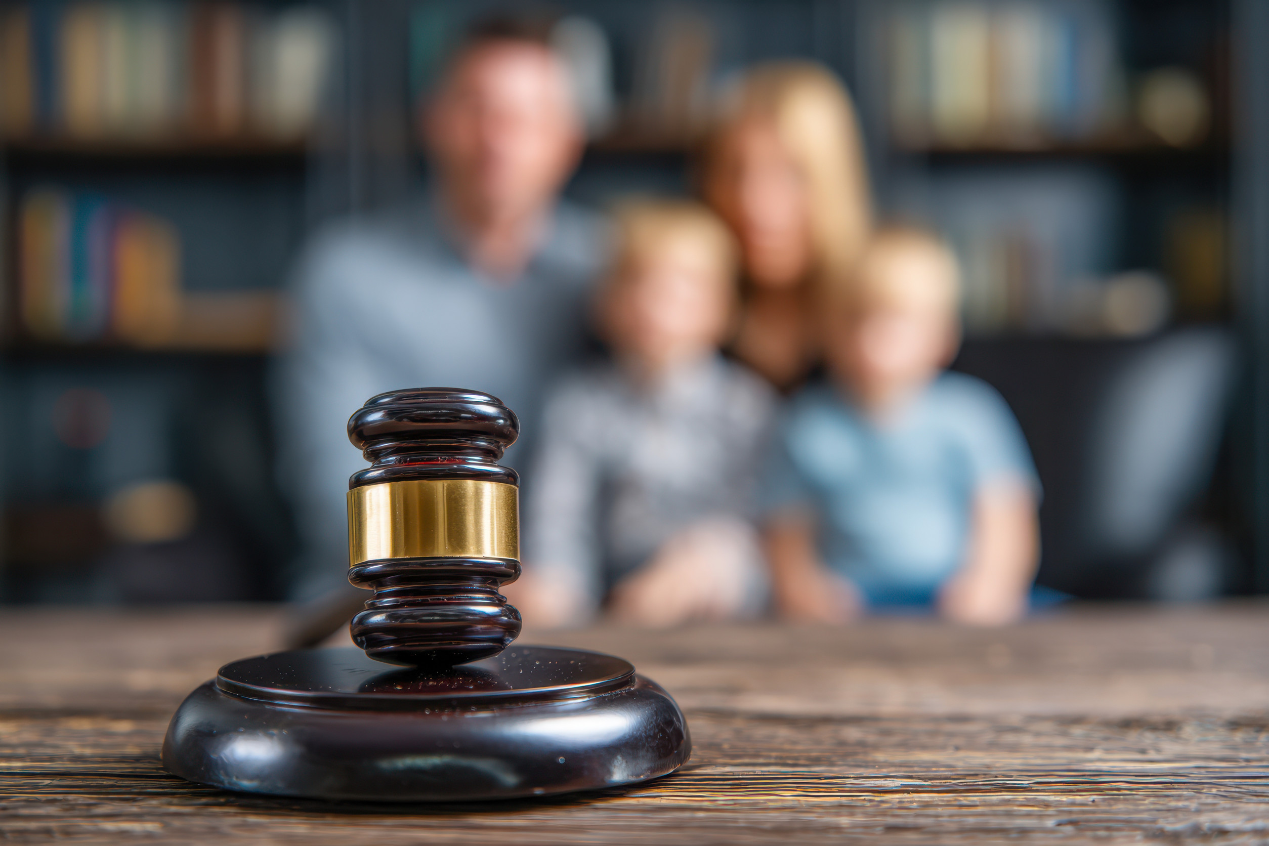 A wooden judge's gavel rests on a table in the foreground, with a blurred family of four sitting together in the background, highlighting a family law or mediation setting designed to be less stressful for families.