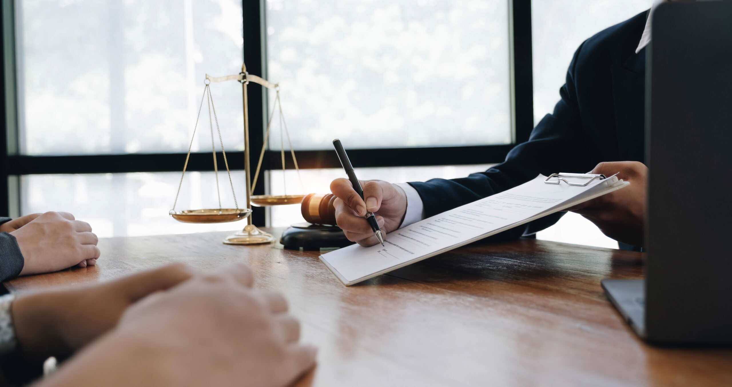 Two people sit across a desk from a person in a suit, who is holding a clipboard and writing. A gavel and legal scales suggest a contract review or consultation—reminding you to carefully review a contract before you sign.