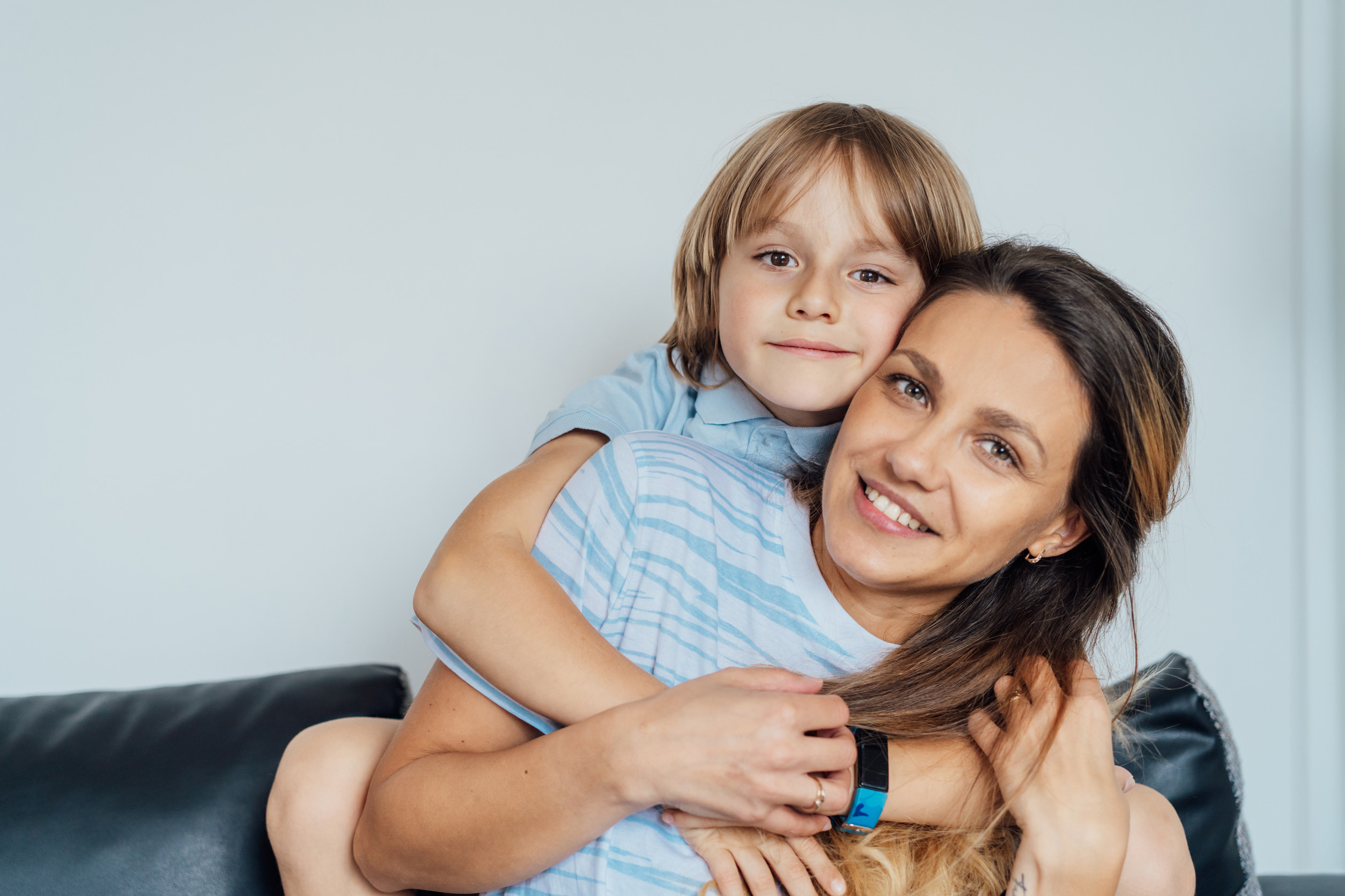 A smiling woman sits on a couch while a young child hugs her from behind, both looking at the camera. They appear happy and relaxed, capturing the joy of parenting time against a plain light background.