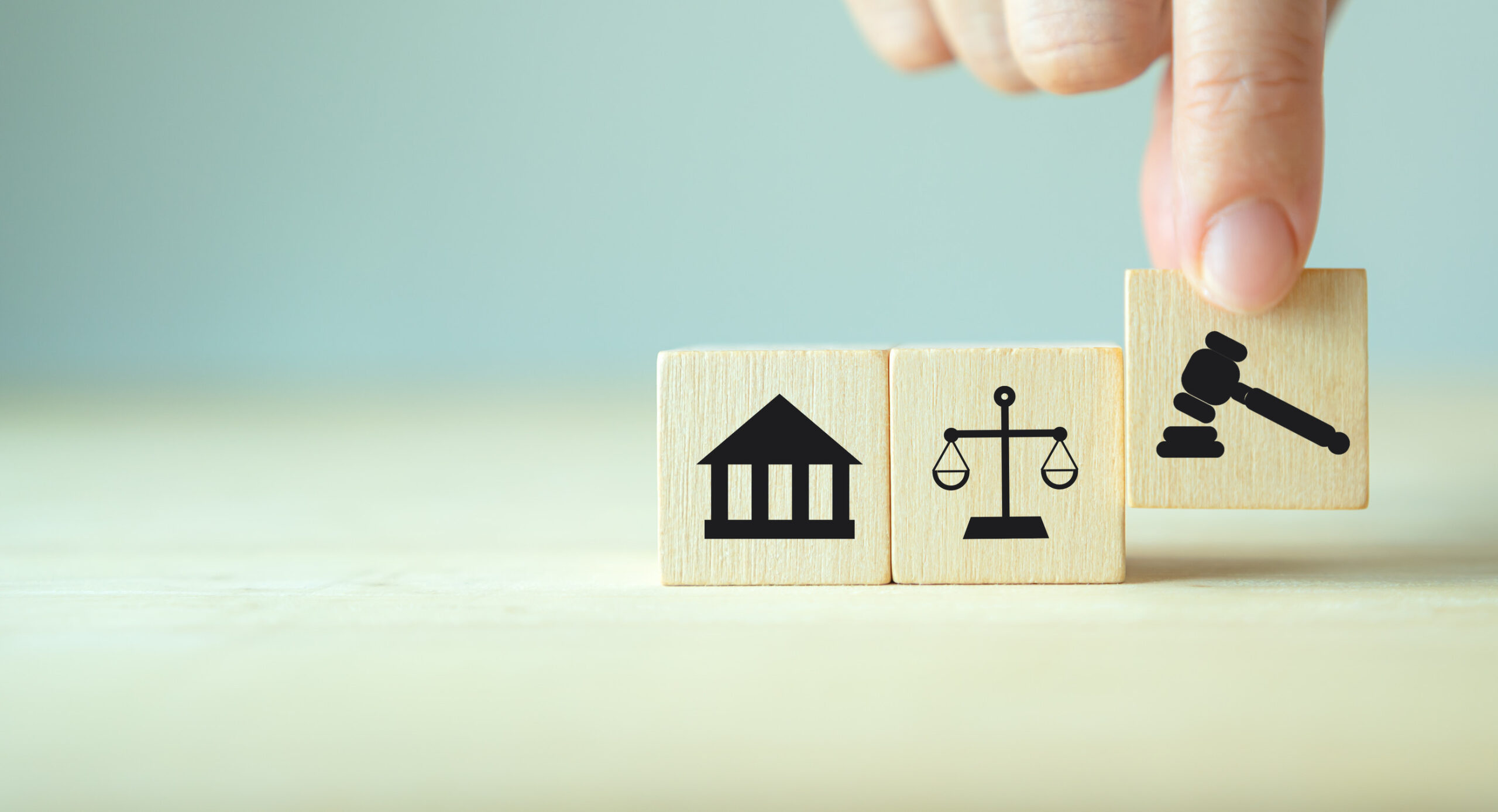 Three wooden blocks on a table show symbols of a courthouse, scales of justice, and a gavel. A hand places the gavel block next to the others, representing legal concepts relevant to Minnesota local government and Open Meeting Law.