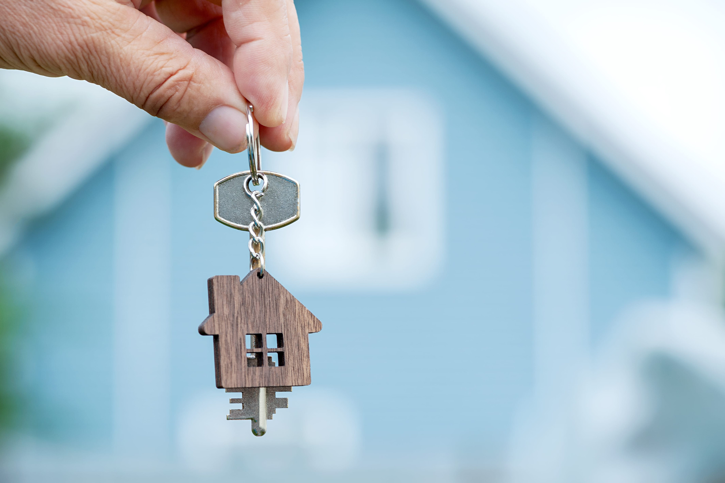 A hand holds a keychain shaped like a house and key in front of a blurred blue house, capturing the excitement of real estate transactions and new purchase agreements.