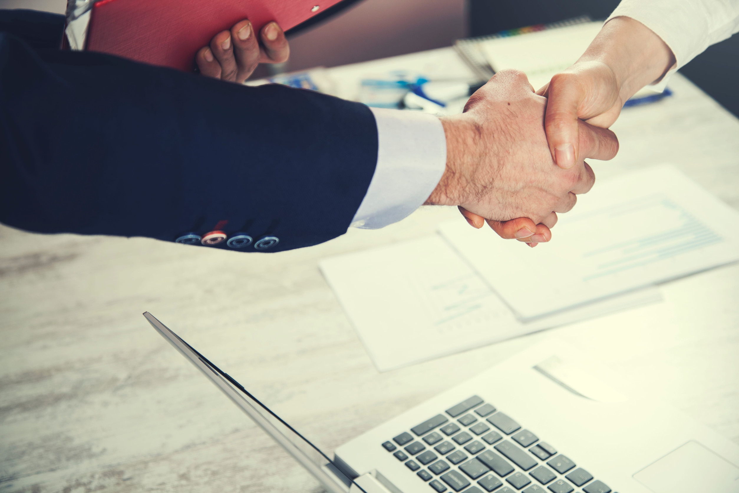 Two people in business attire shaking hands over a desk with a laptop, documents, and a red folder visible, suggesting a successful meeting or agreement in a professional office setting.