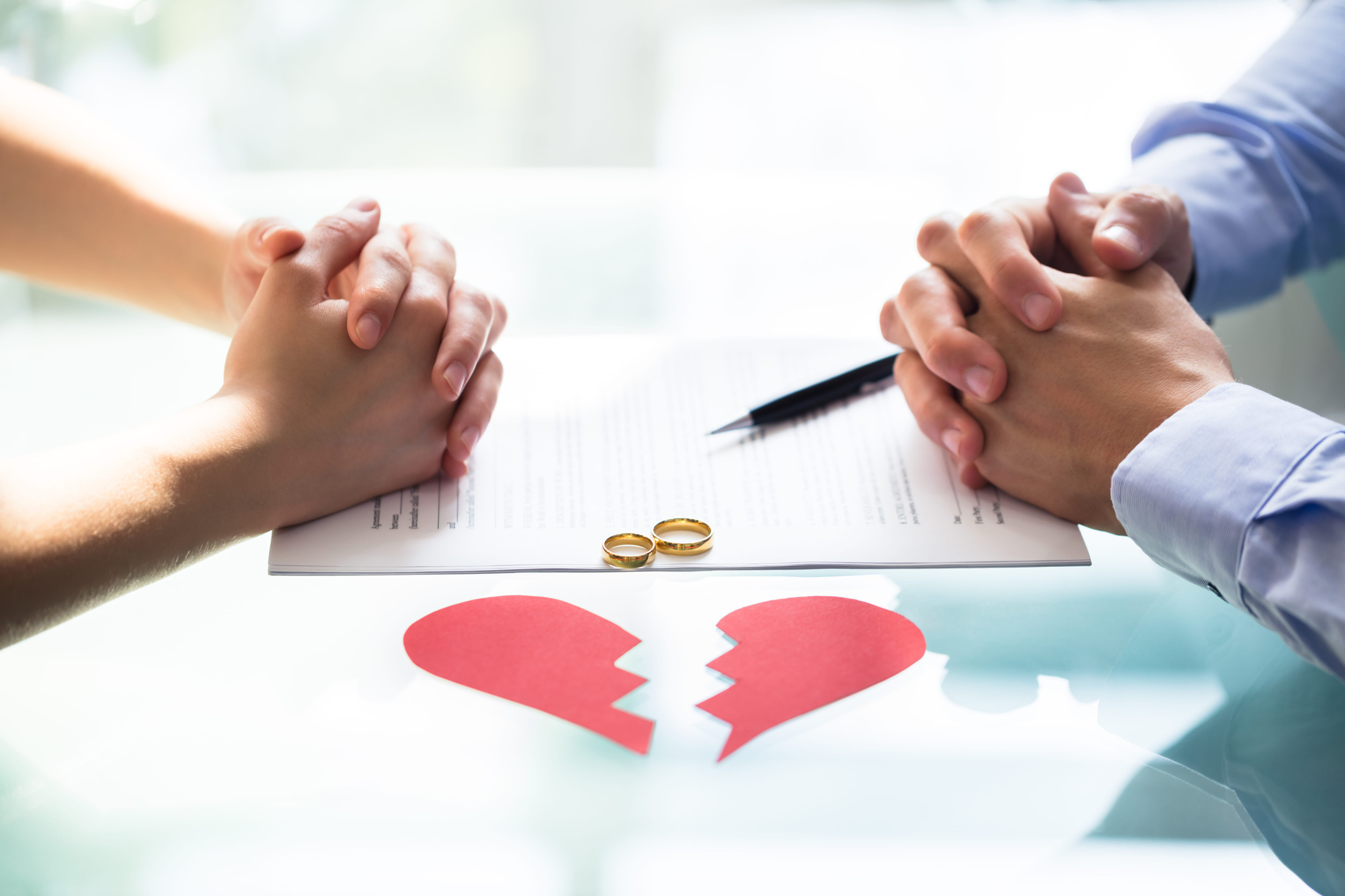 Two people sit across from each other with hands clasped over a document, two wedding rings, and a red broken heart cutout, symbolizing the Minnesota divorce process or separation.