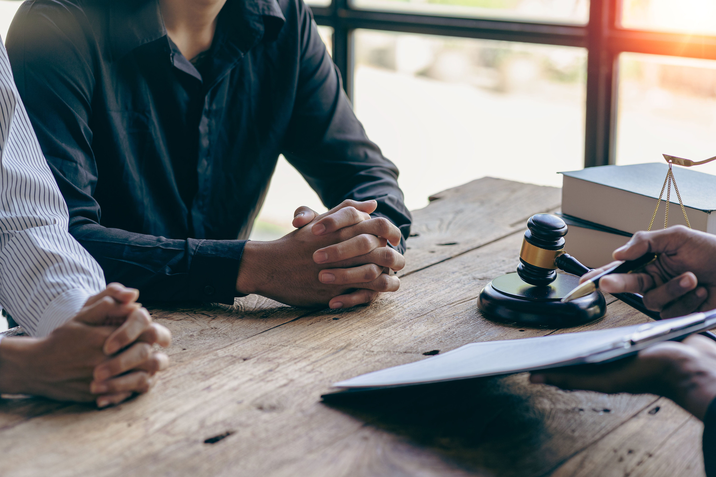 Three people sit at a wooden table with hands clasped; one holds a clipboard, while a gavel and legal books rest nearby, illustrating what mediation looks like during the mediation process or a consultation meeting.
