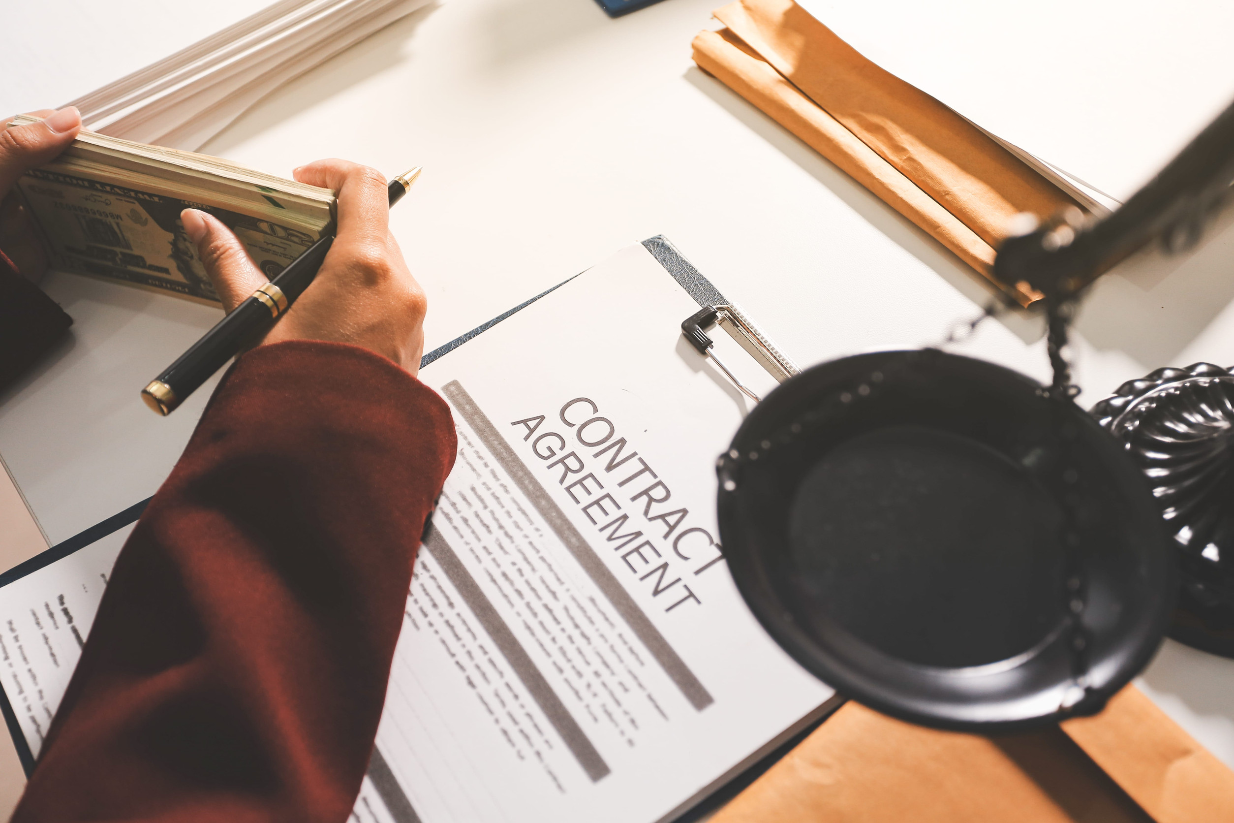 A person holds a stack of cash and a pen near a clipboard with a contract agreement document on a desk, representing government operations, alongside legal scales and folders.