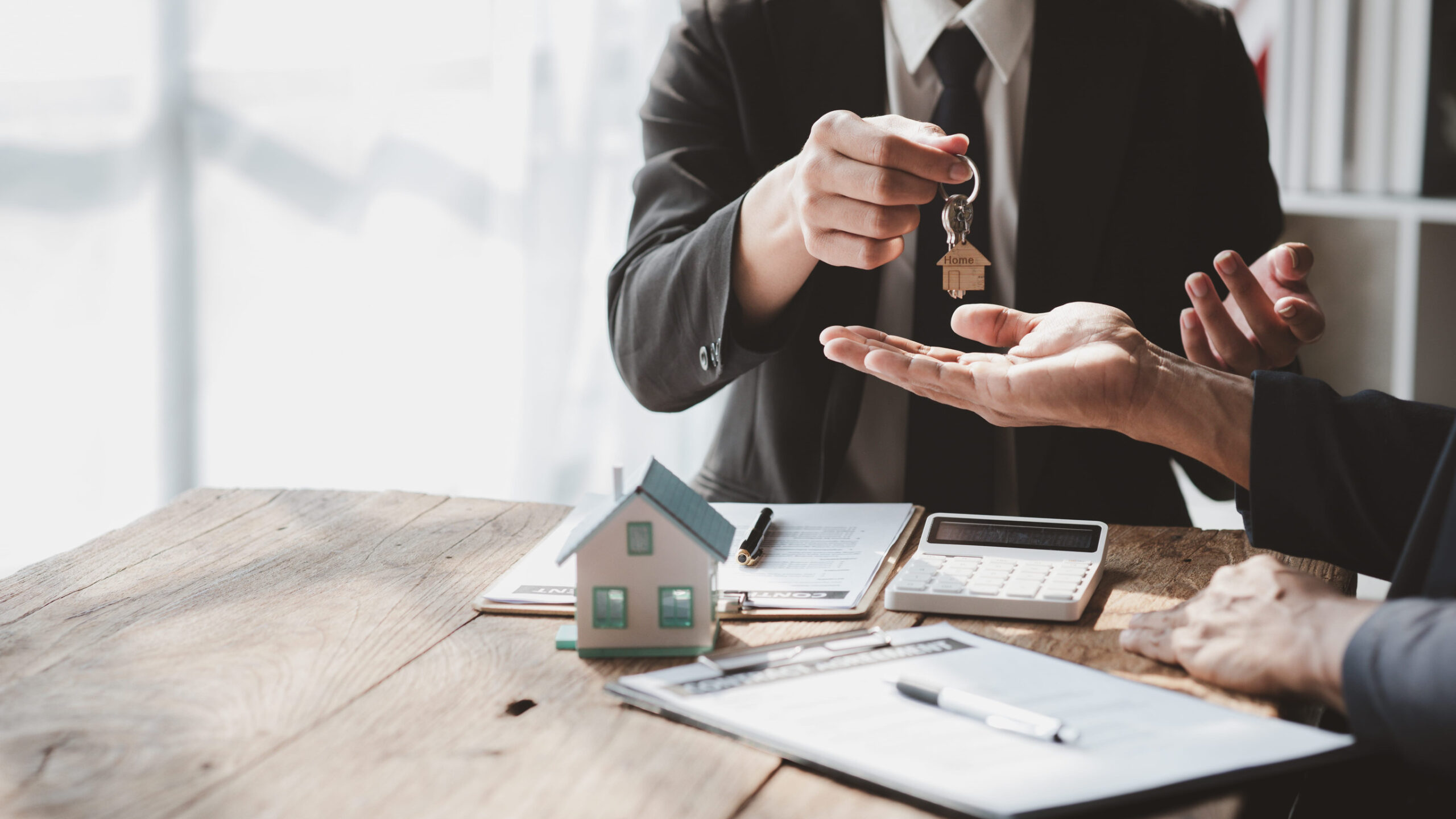 A person in a suit hands over a house key to another person across a wooden table with documents, a calculator, and a small model house, depicting what to expect at closing during the real estate closing process.