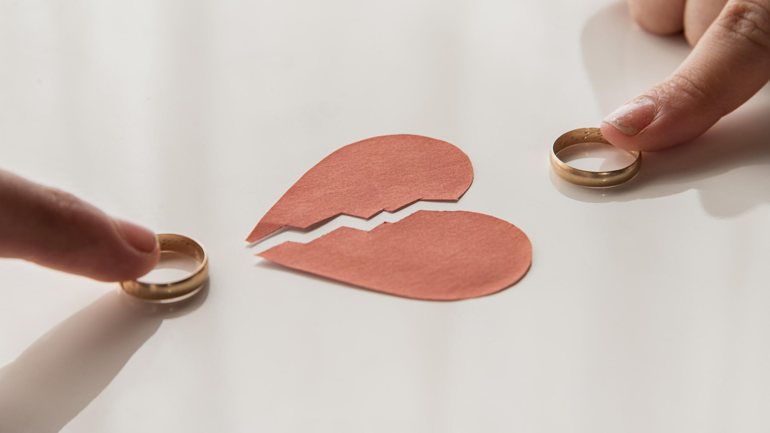 Two hands each hold a wedding ring, placing them beside a broken paper heart on a white surface, symbolizing divorce in Minnesota or separation.