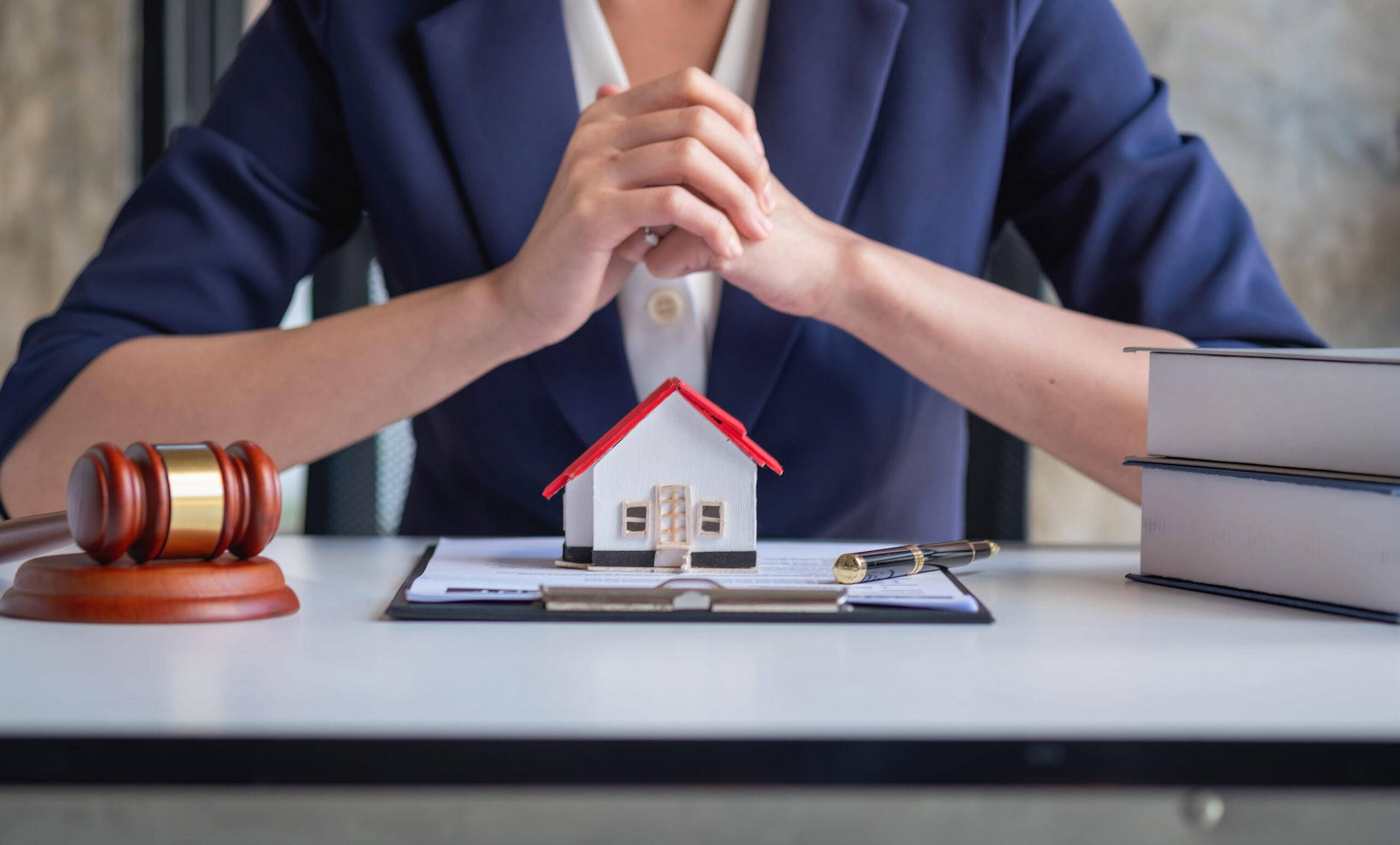 A person in a suit sits at a desk with hands clasped over a small model house, surrounded by a gavel, pen, and legal documents, suggesting issues like inheritance in Minnesota or what happens if you die without a will in Minnesota.