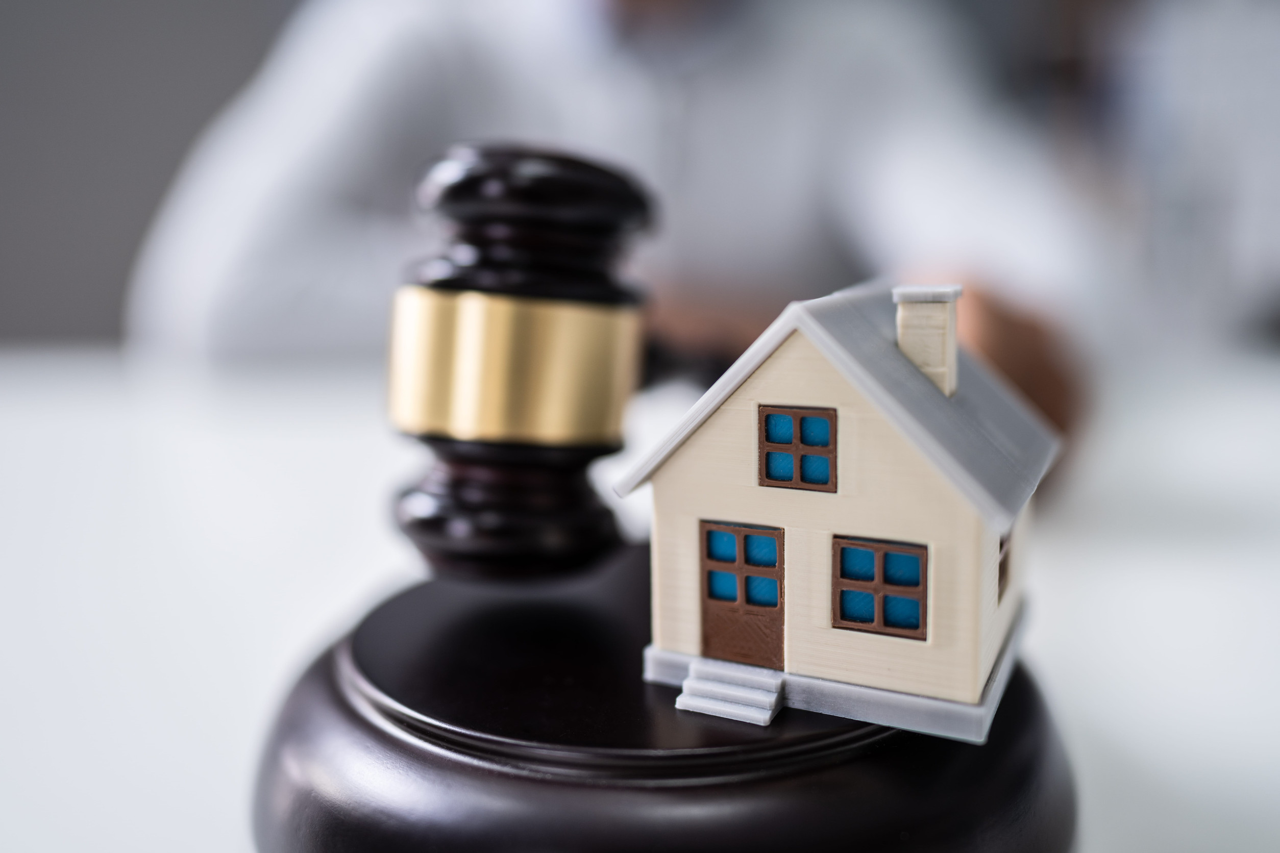 A small model house sits next to a judge’s gavel on a desk, symbolizing real estate law or property disputes. This scene reflects the vital role of a Minnesota real estate attorney during a real estate closing in Minnesota. A blurred person is visible in the background.
