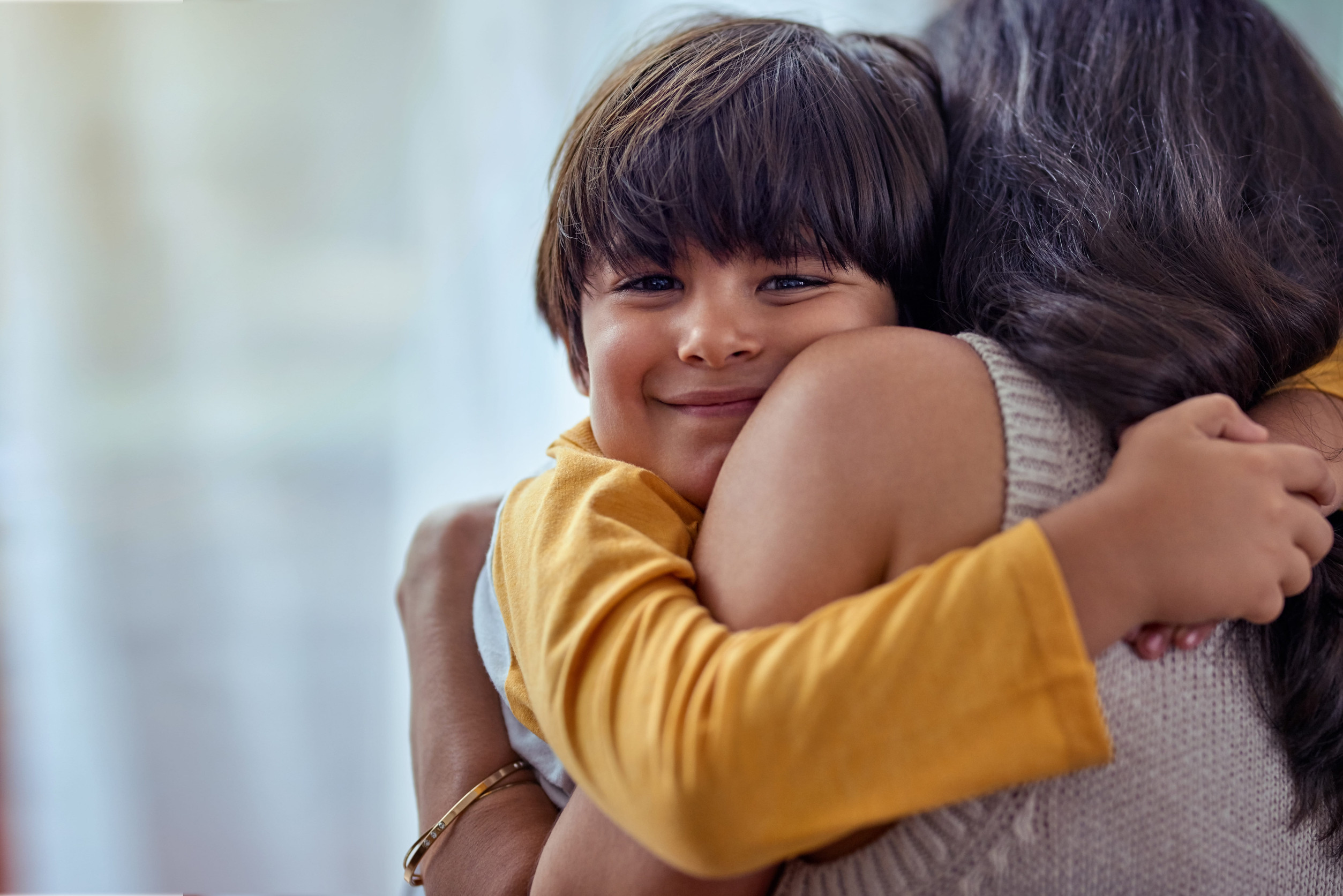 A young boy with brown hair and a yellow shirt smiles while hugging a woman with long dark hair, whose back is to the camera, in a warm, close embrace—capturing precious Parenting Time Minnesota moments indoors.
