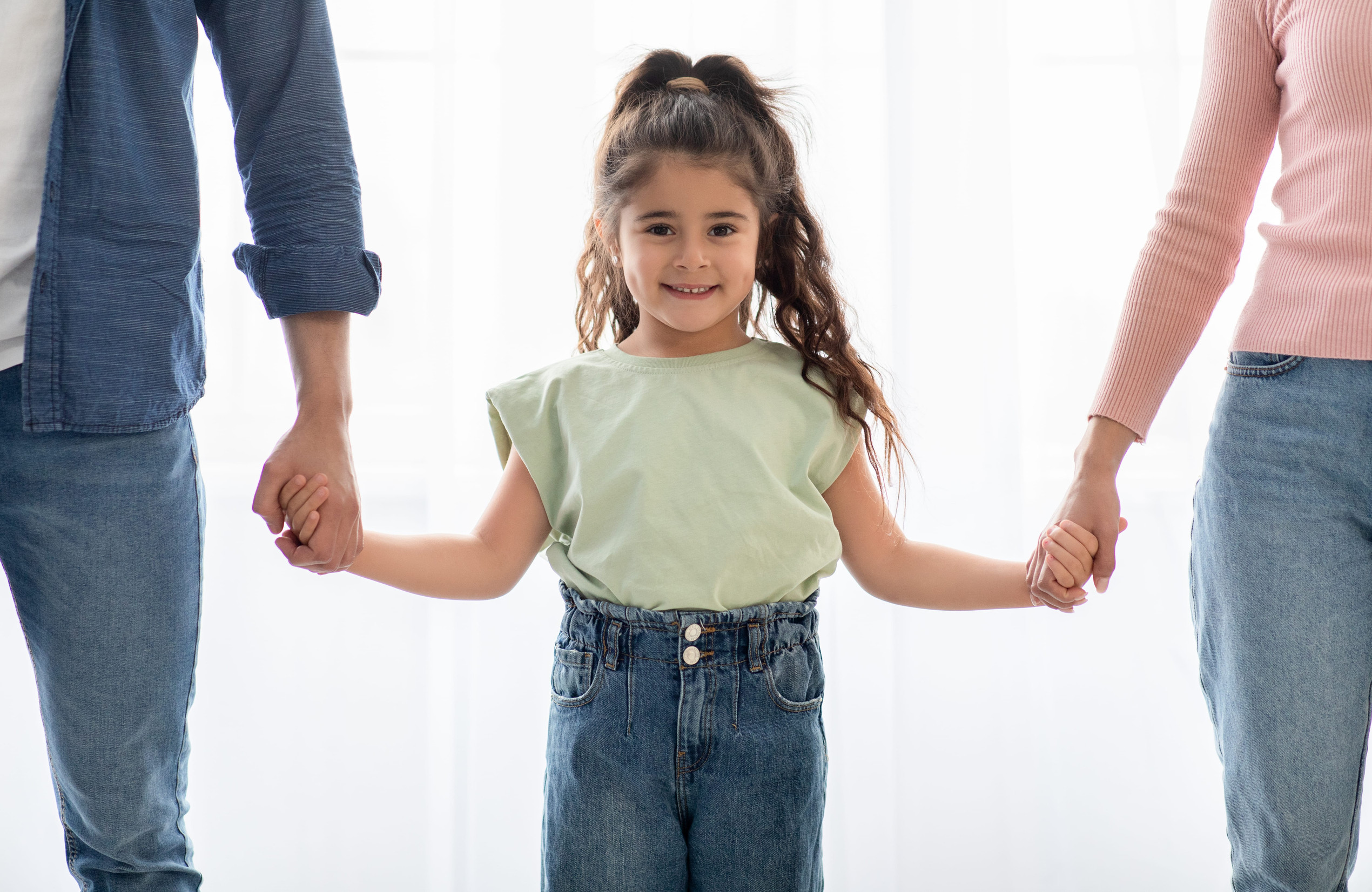 A young girl with long hair stands smiling, holding hands with two adults—perhaps navigating parenting after divorce in Minnesota. The adults are partially visible, dressed casually against a bright, softly lit background.