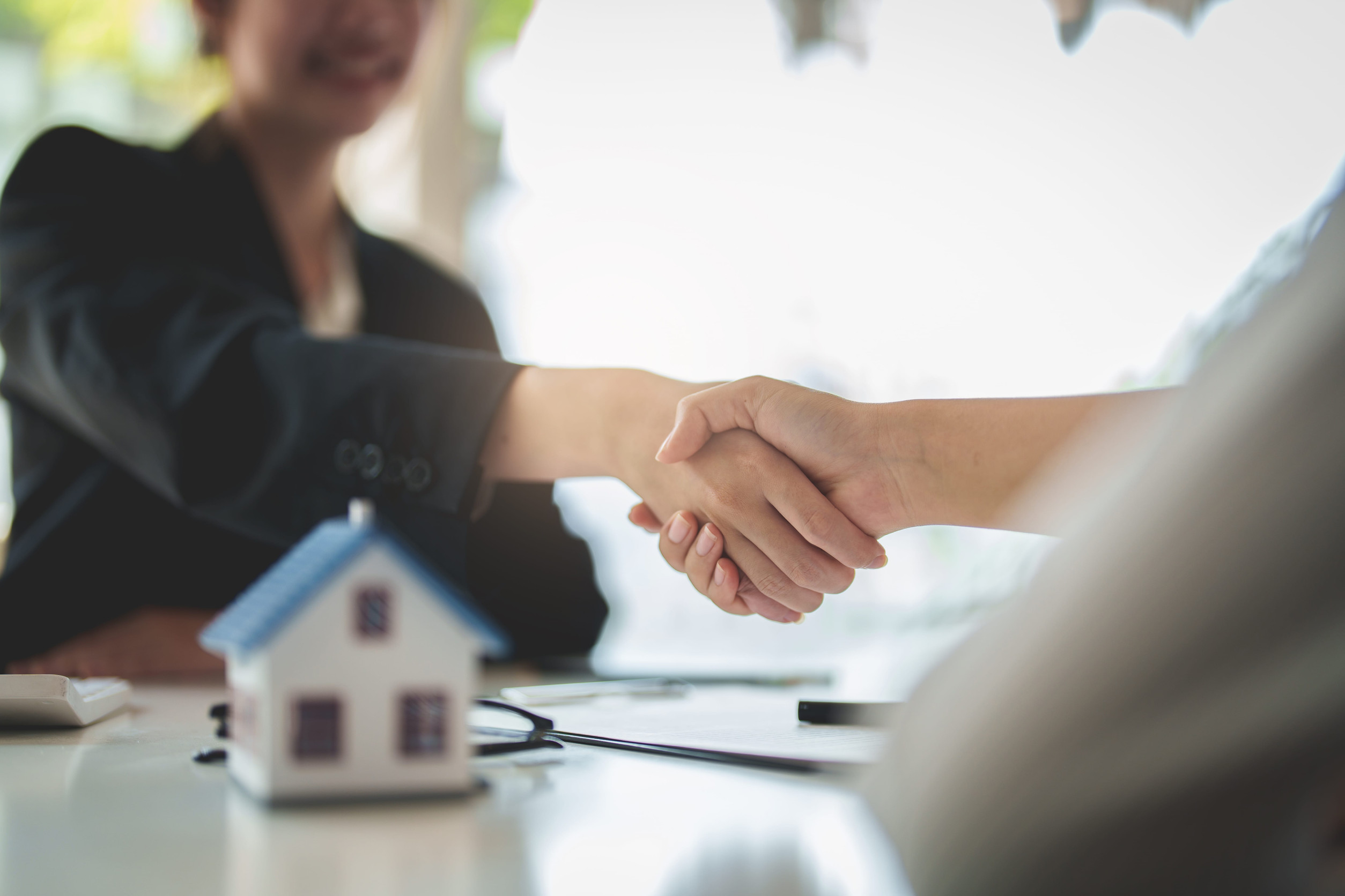 Two professionals shake hands over a desk with a small house model, signaling a real estate deal or partnership.
