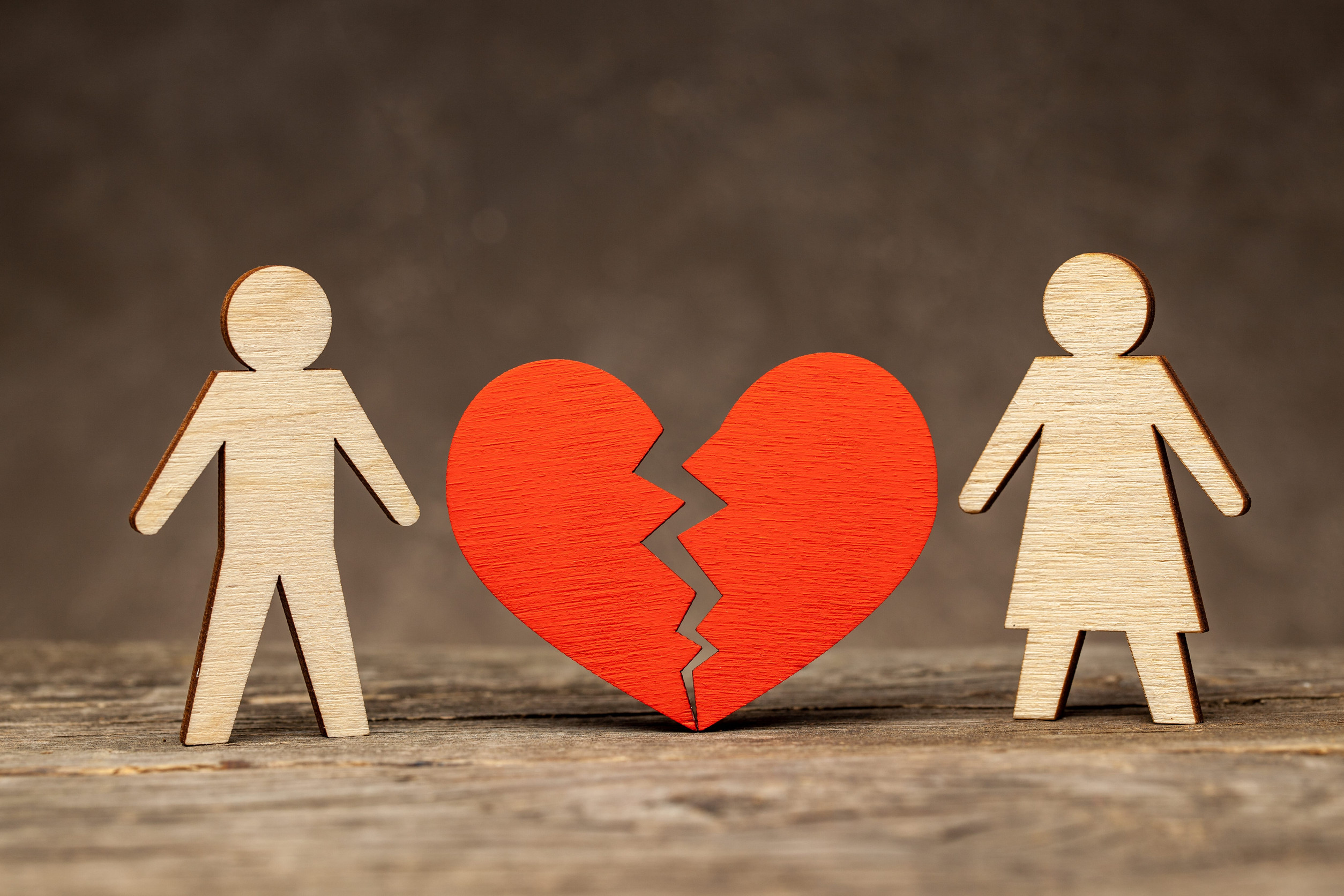 Two wooden figures, one male and one female, stand on either side of a red broken heart on a wooden surface, symbolizing separation or heartbreak—much like the emotional distance that can occur during a Legal Separation in Minnesota or Divorce in Minnesota.