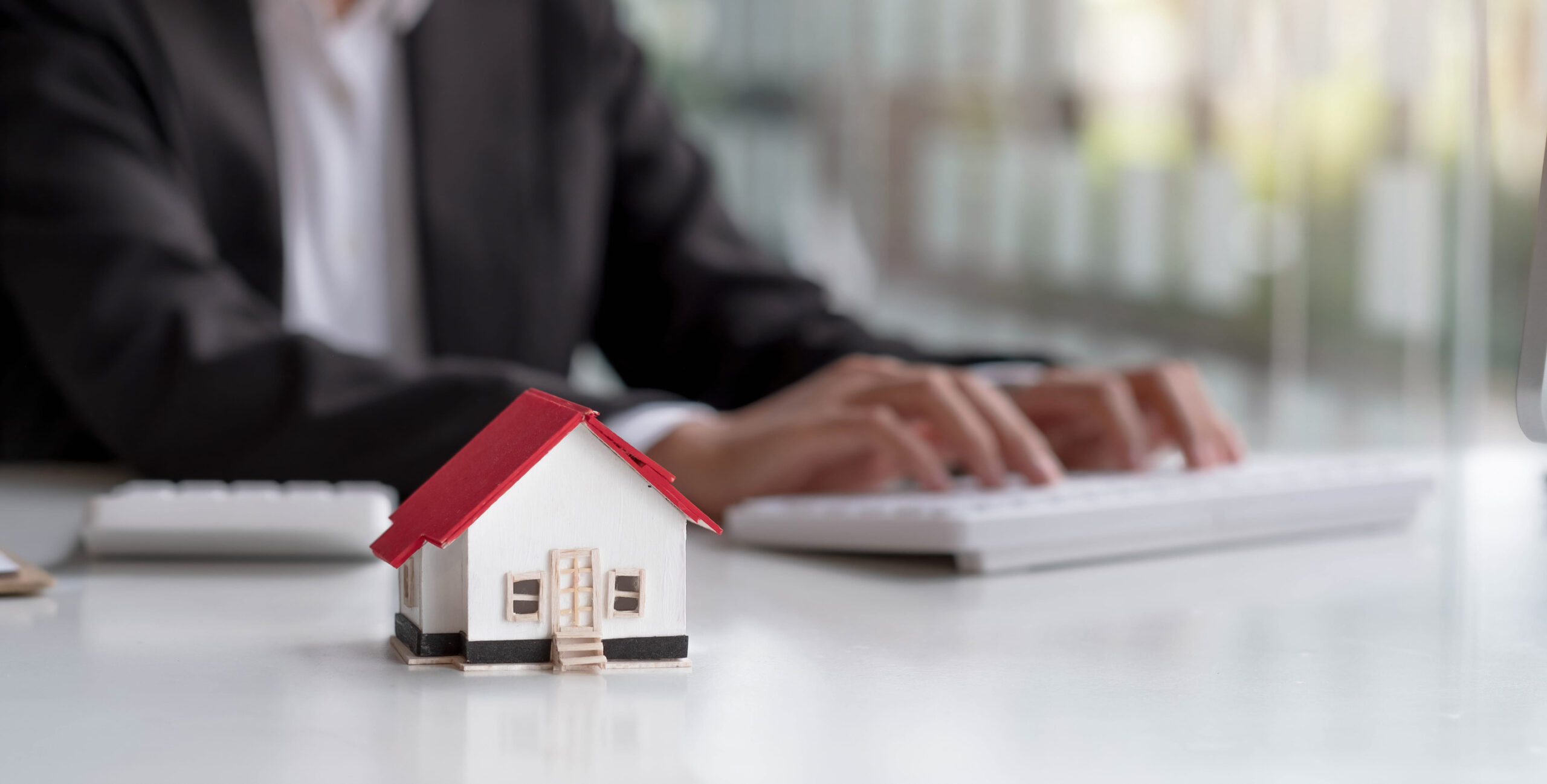 A small model house with a red roof sits on a desk, while a person in a suit types on a keyboard in the blurred background—evoking the meticulous nature of Estate Administration Minnesota professionals as they navigate Minnesota estate laws.