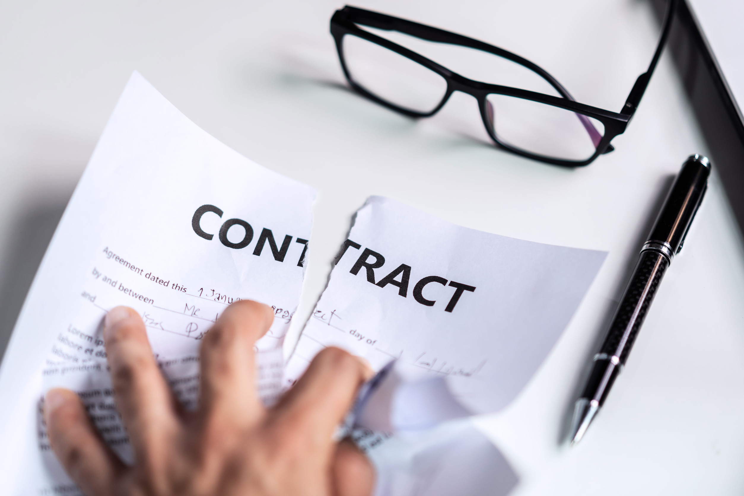 A hand tearing a contract in half on a white desk, with eyeglasses and a black pen nearby, depicting a breach of contract scenario in Minnesota.