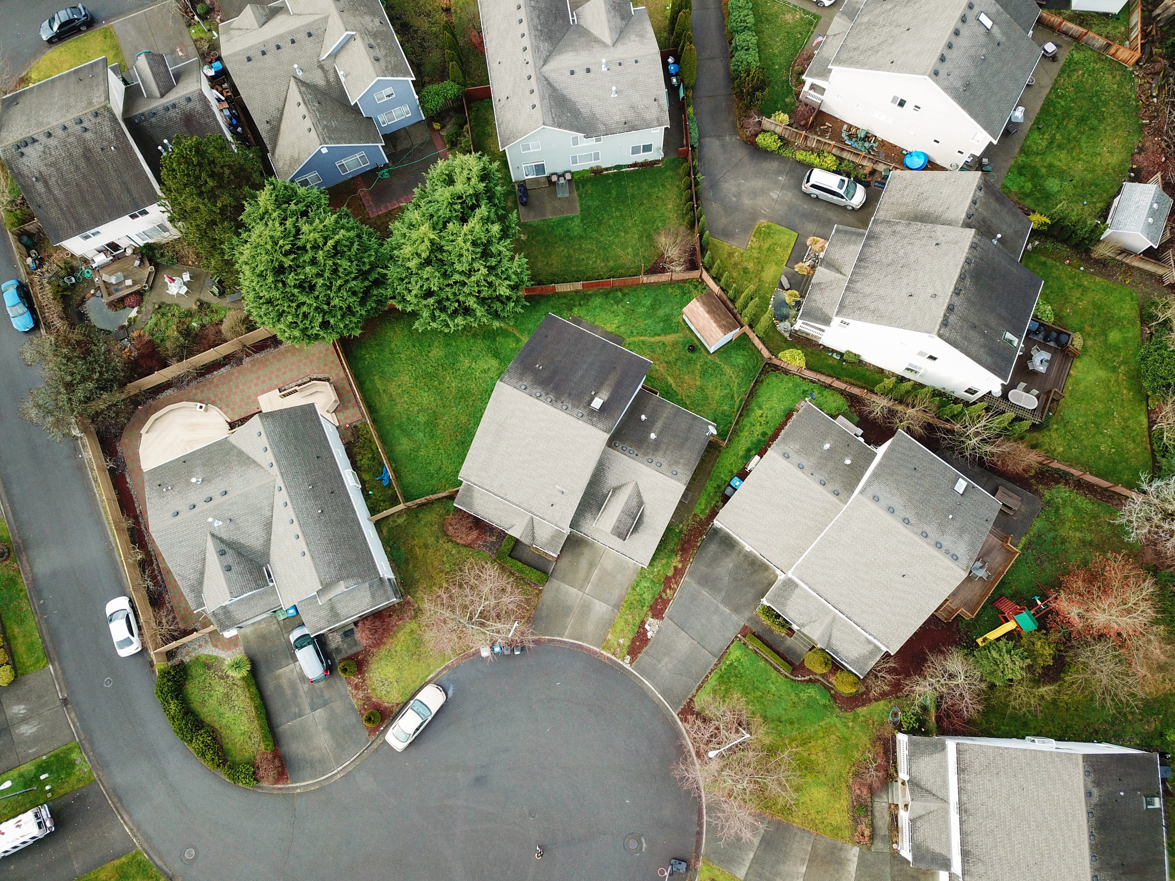 Aerial view of suburban houses with gray roofs, driveways, green lawns, and trees in Minnesota. Cars are parked along the curved street, and fenced yards separate each home, reflecting clear property rights and visible easements between properties.